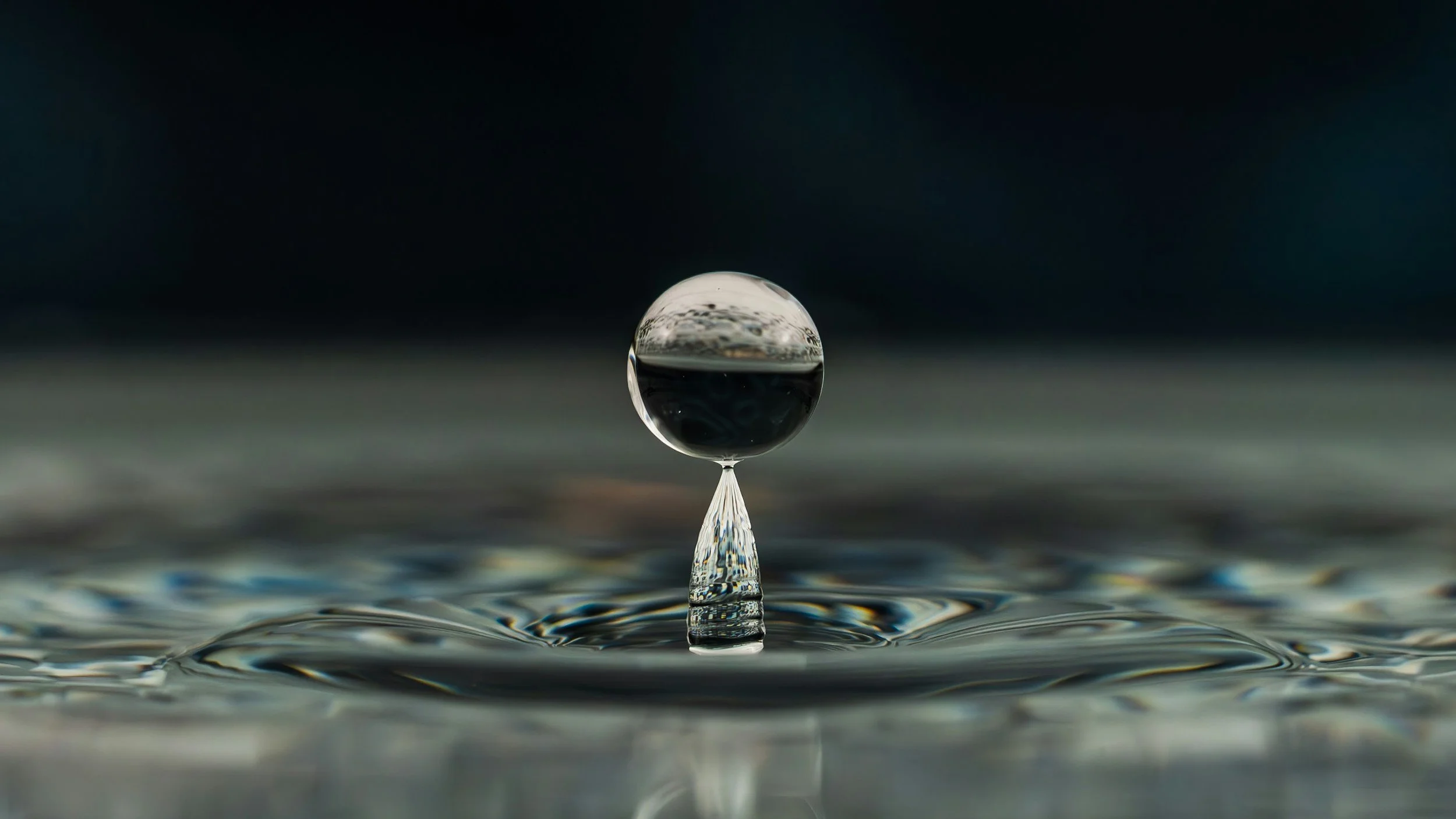Close-up of a single water droplet balanced on a thin stem, with a reflective surface below and a dark background.