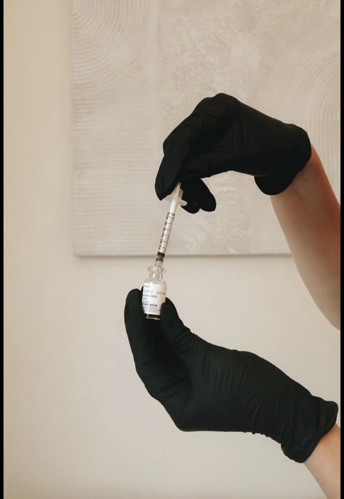 A person wearing black gloves holding a syringe and vial in front of a light-colored textured wall.