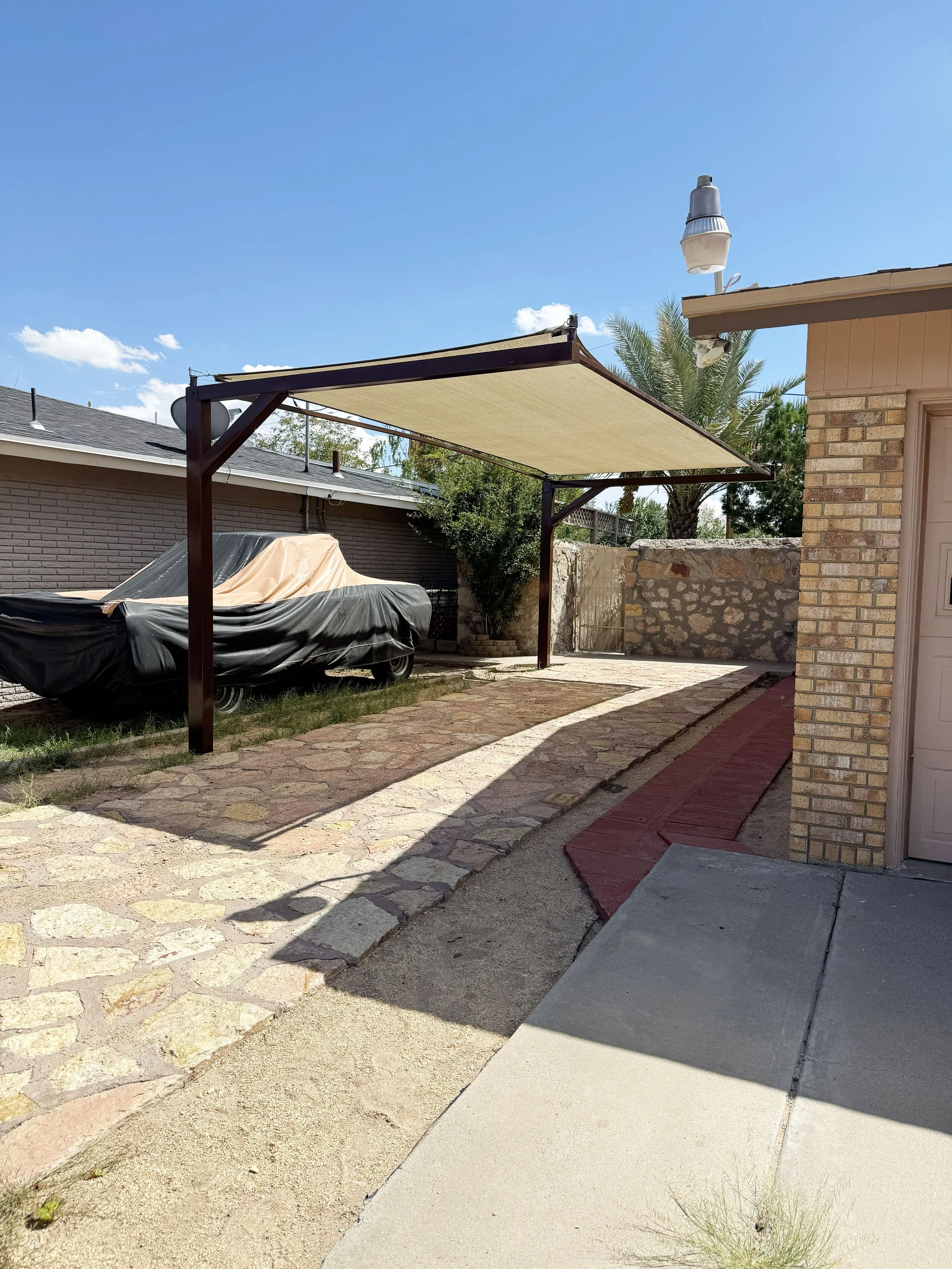 A residential backyard with a covered carport, a boat covered with a tarp, and a brick house with outdoor lighting fixtures under a clear blue sky.