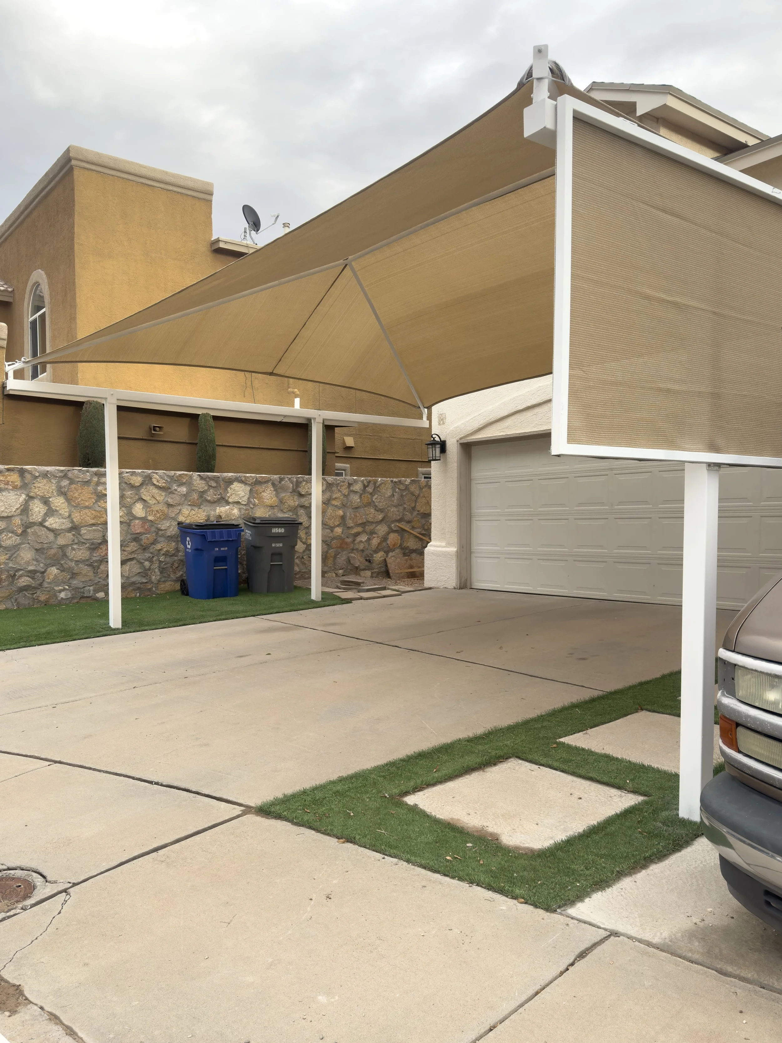 A residential house with a garage, a driveway, and two trash bins. There is a beige retractable awning over the driveway and a small strip of artificial grass near the edge of the driveway.