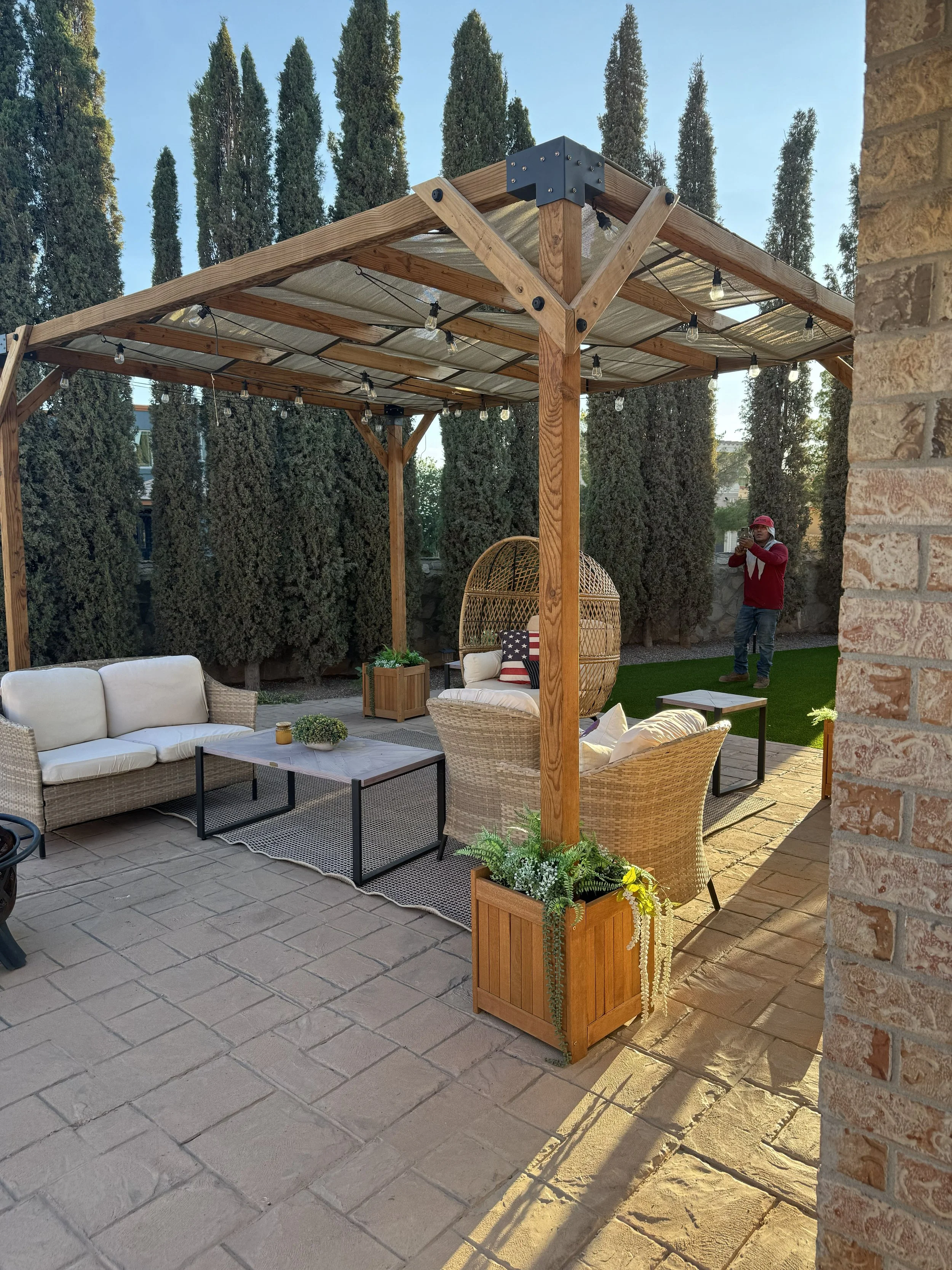 A backyard patio with outdoor furniture, a wooden pergola with string lights, potted plants, and a person in the background taking a photo.