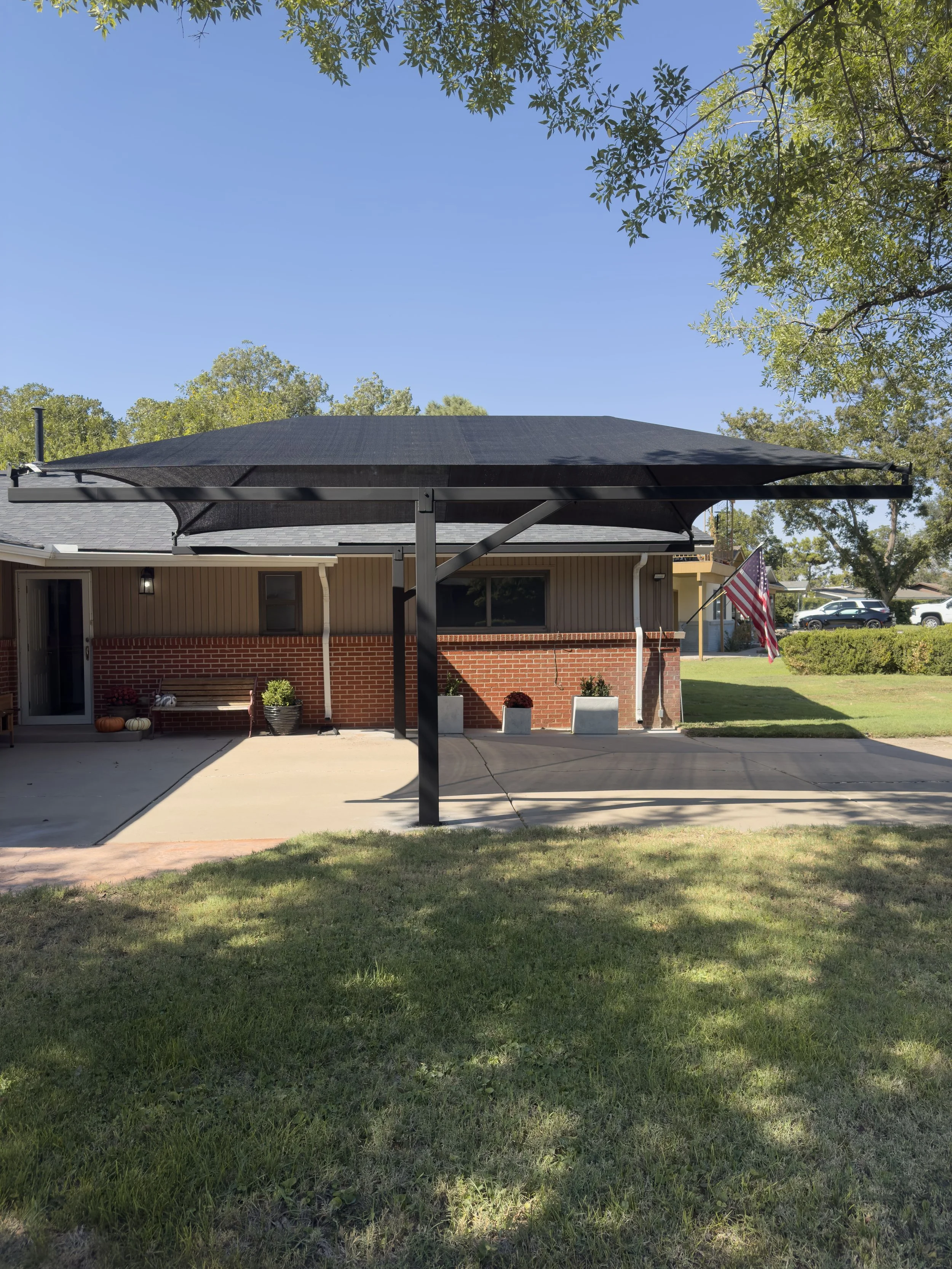 A backyard patio with a black sunshade canopy, a bench, potted plants, pumpkins, and an American flag on a pole, with a house in the background and a clear blue sky.