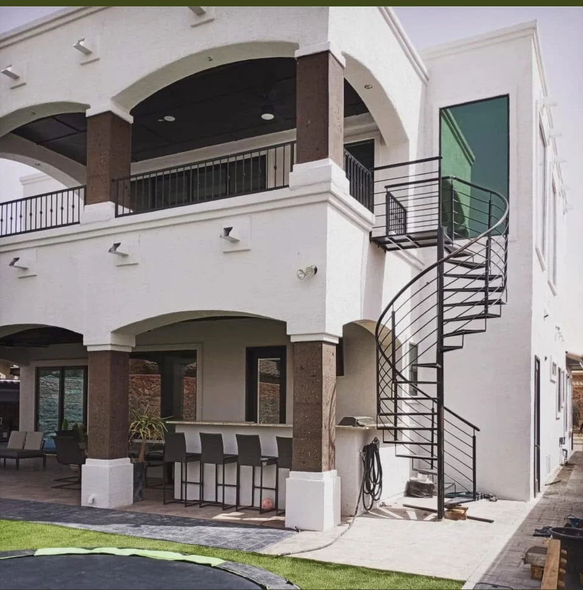 The exterior of a multi-story house with white walls, brown columns, and a spiral staircase on the side, leading to the upper balcony.