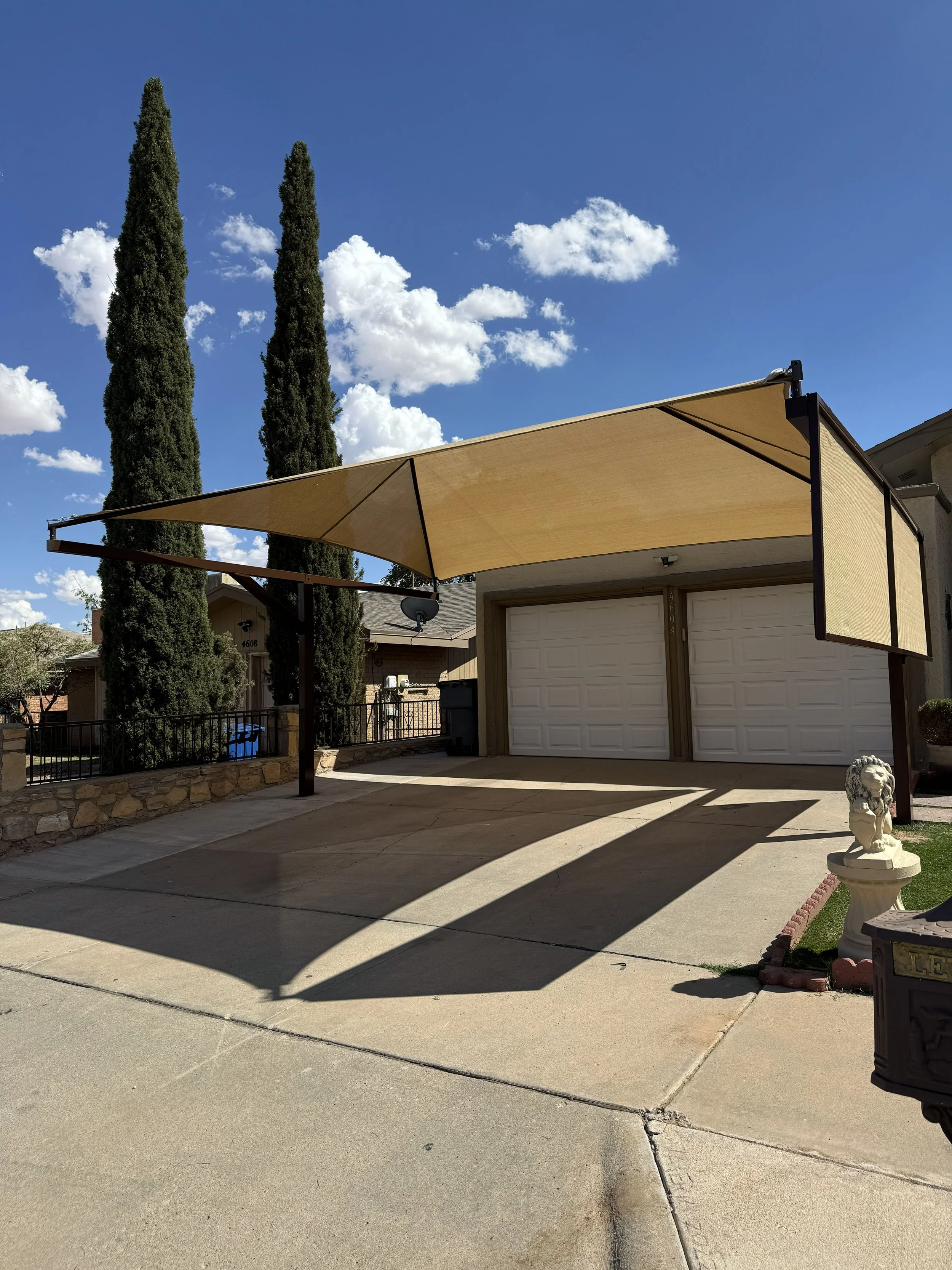 Residential driveway with a beige canopy providing shade, white garage doors, and tall cypress trees against a blue sky with scattered clouds.