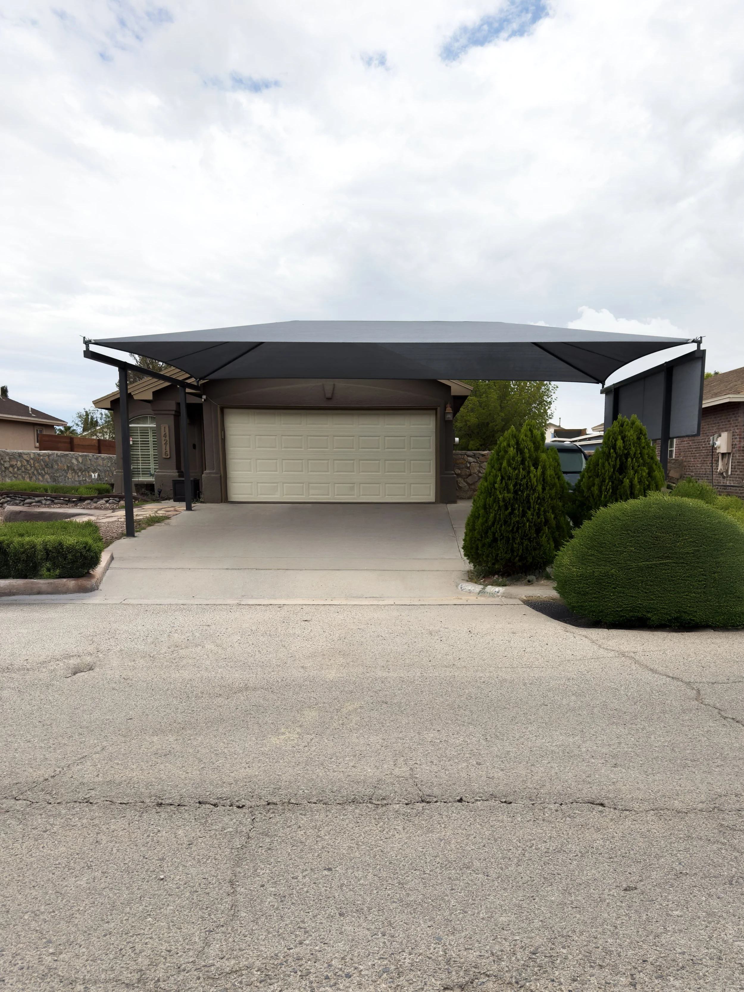 Front view of a house with a concrete driveway, a closed white garage door, and a black retractable awning above the driveway, with bushes and trees on the yard.