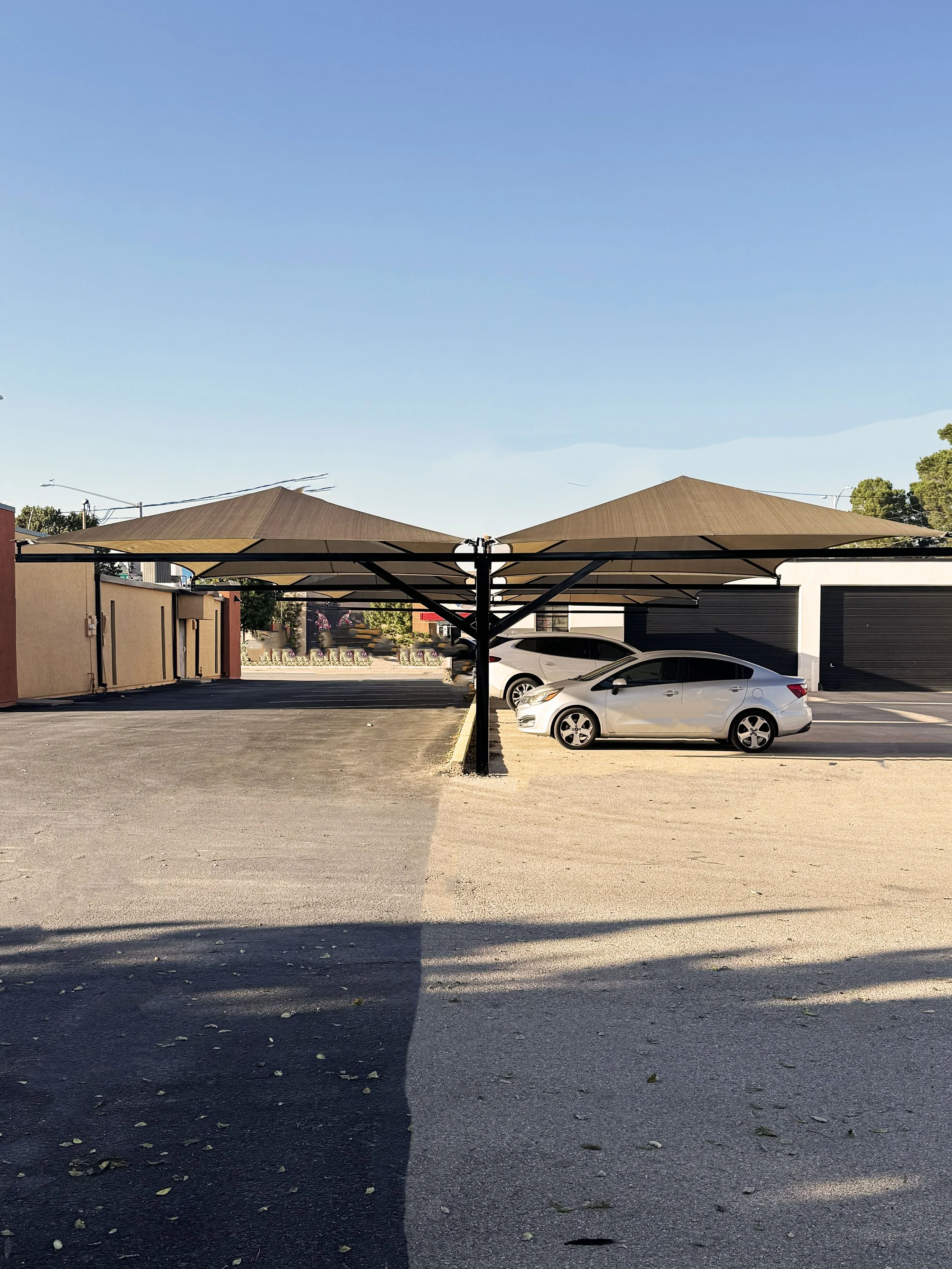 Outdoor parking lot with two beige shade sails, a white car parked under one shade, and a black garage door in the background under a clear blue sky.