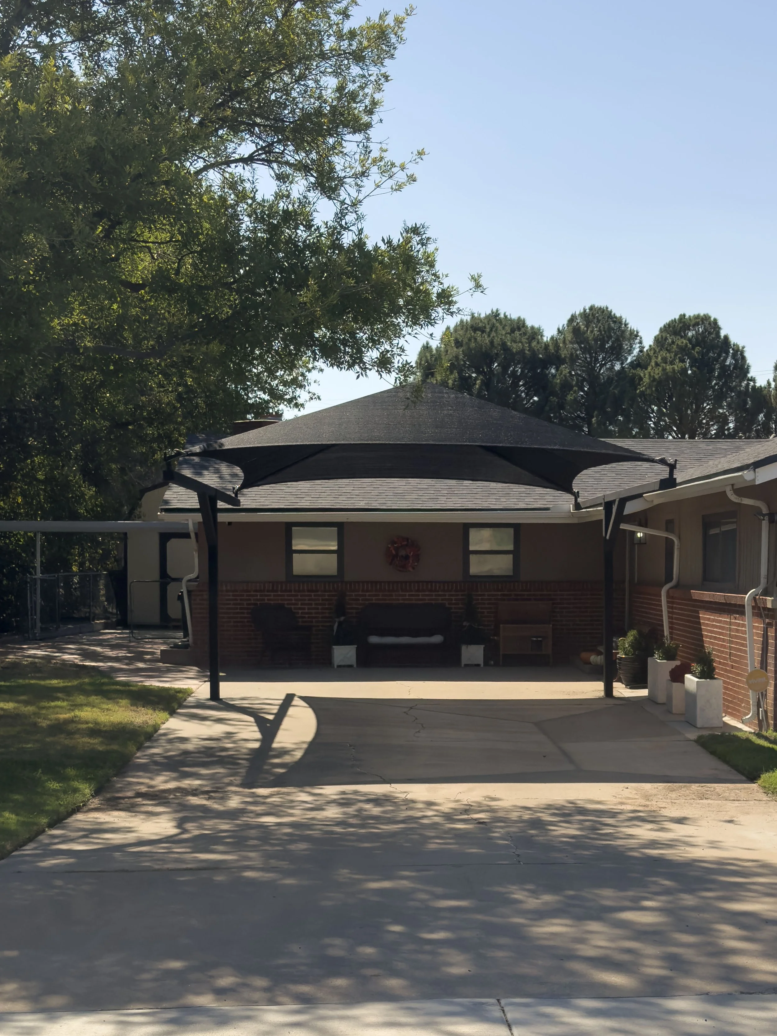 A residential house with a concrete driveway, a shaded porch area with outdoor seating, a black canopy, potted plants, and trees in the background.