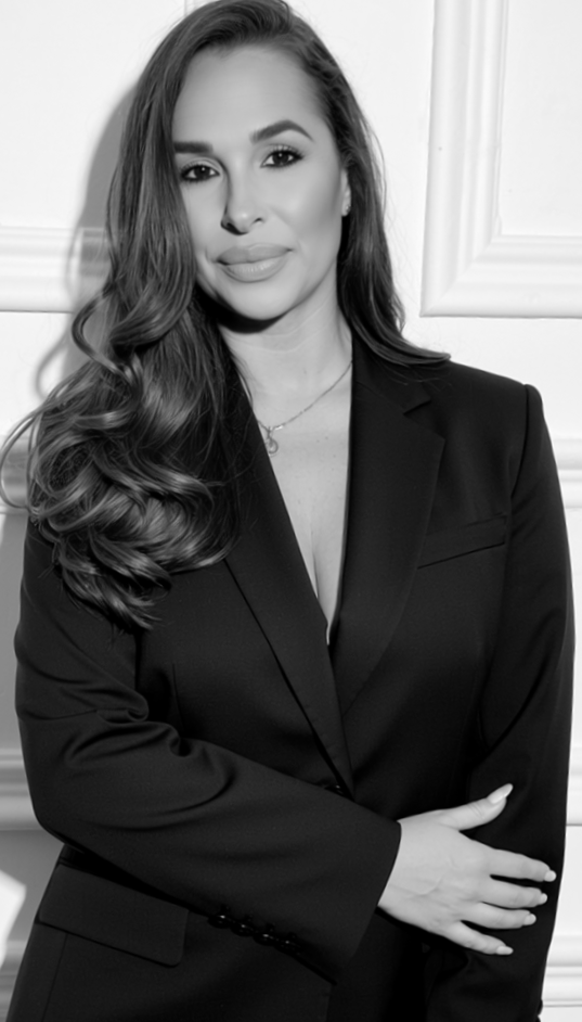 Black and white photo of a woman in a blazer with long, wavy hair, standing in front of a white panel wall. She is looking at the camera with a slight smile, arms crossed.