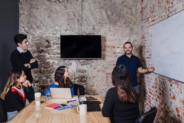 Group of five people in a business meeting in a rustic office, with one person presenting near a whiteboard.