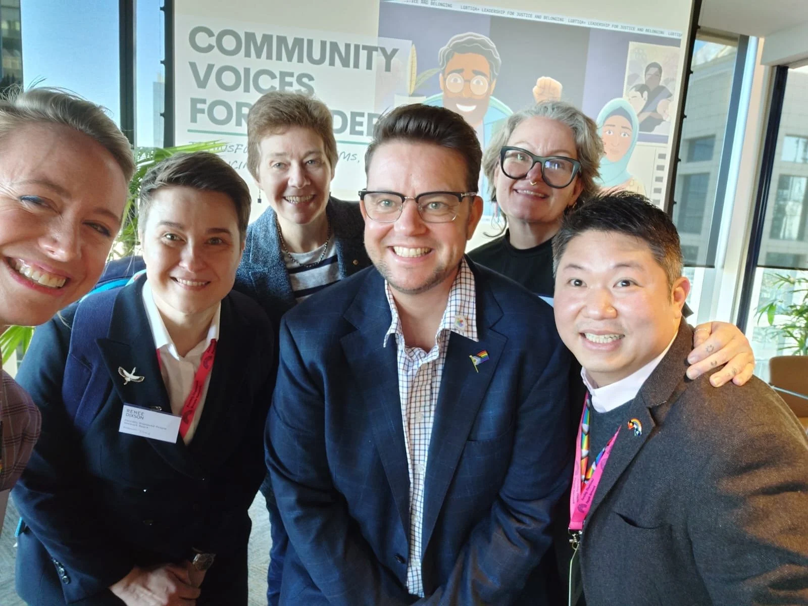 Group of LGBTQIA+ community and sector leaders smiling together at a community event in front of a “Community Voices” banner.