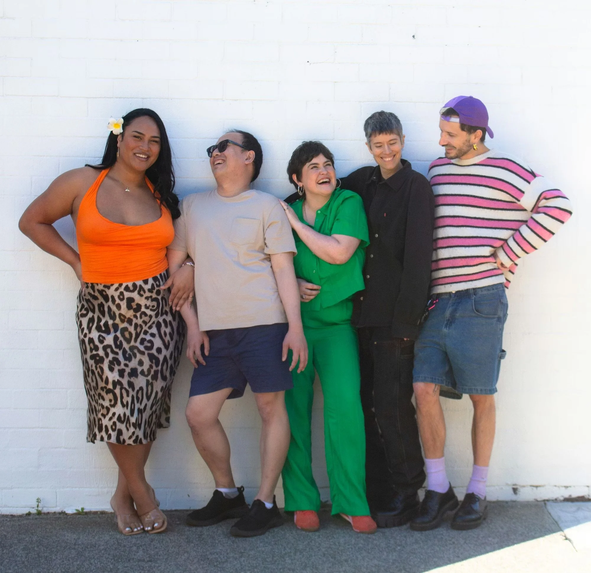 Five LGBTQIA+ community members standing together laughing and smiling against a white wall in bright, colourful casual clothing.