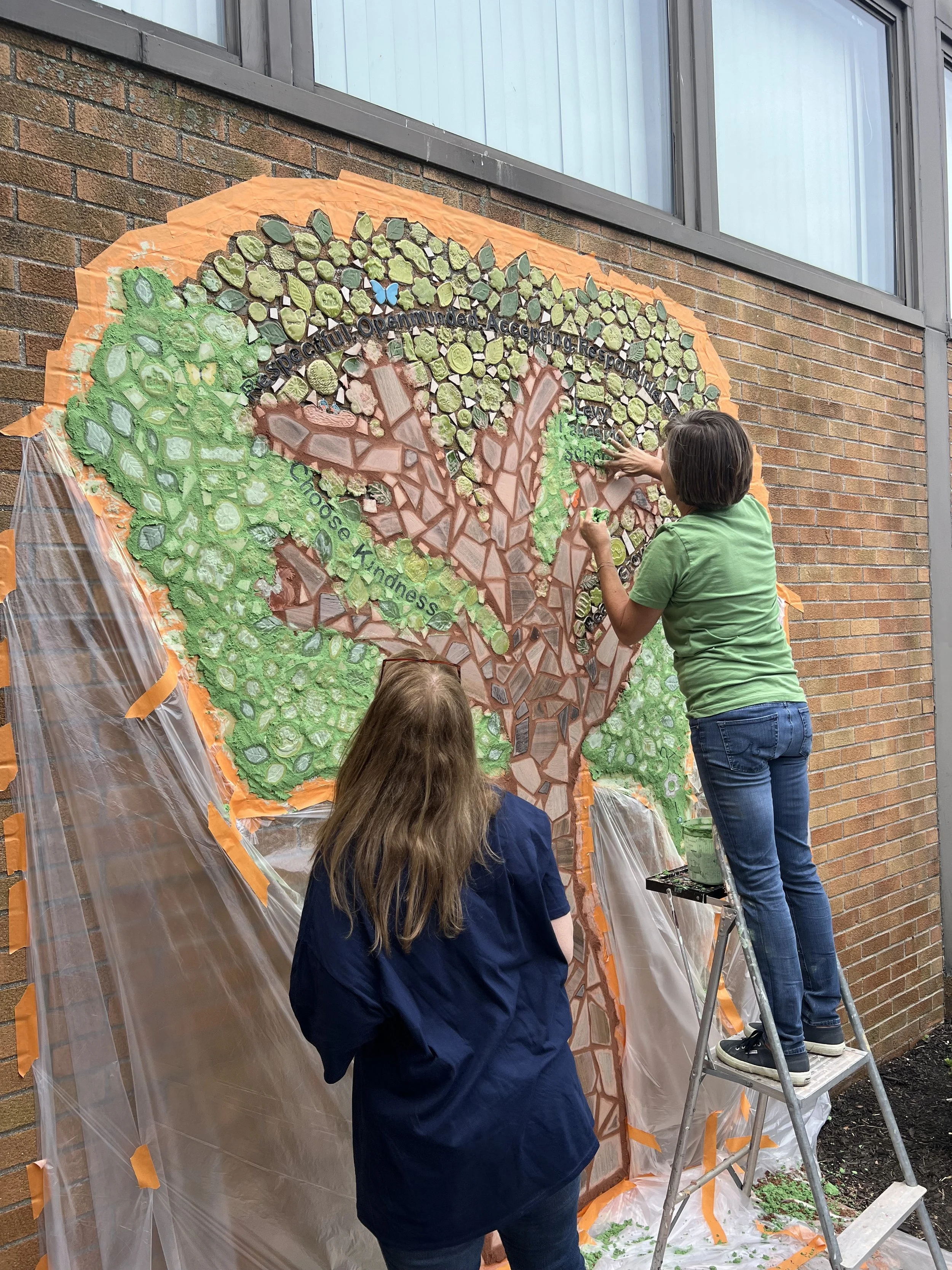 Two women working on a large outdoor mosaic wall art of a tree, with one standing on a ladder and the other observing.