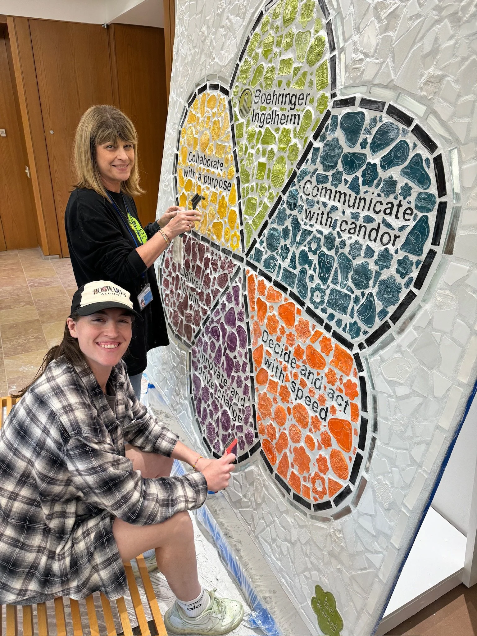 Two women working on a colorful mosaic wall art with inspirational words and symbols. One woman is standing, and the other is kneeling with a smile, both engaging with the artwork.