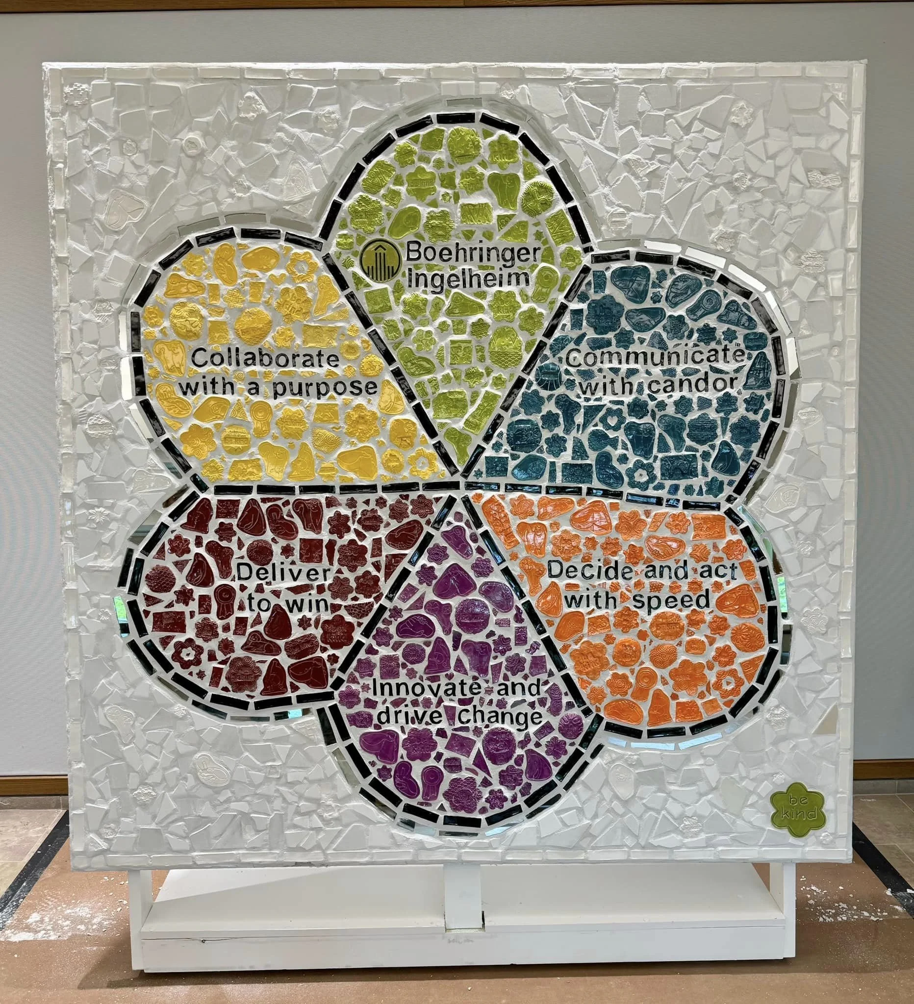 A colorful mosaic art piece with five overlapping petals, each containing text and icons. The petals are labeled: 'Boehringer Ingelheim,' 'Communicate with candor,' 'Decide and act with speed,' 'Innovate and drive change,' and 'Deliver to win.' The background is white with small, textured pieces, and there is a small green plaque at the bottom right that says "be kind."
