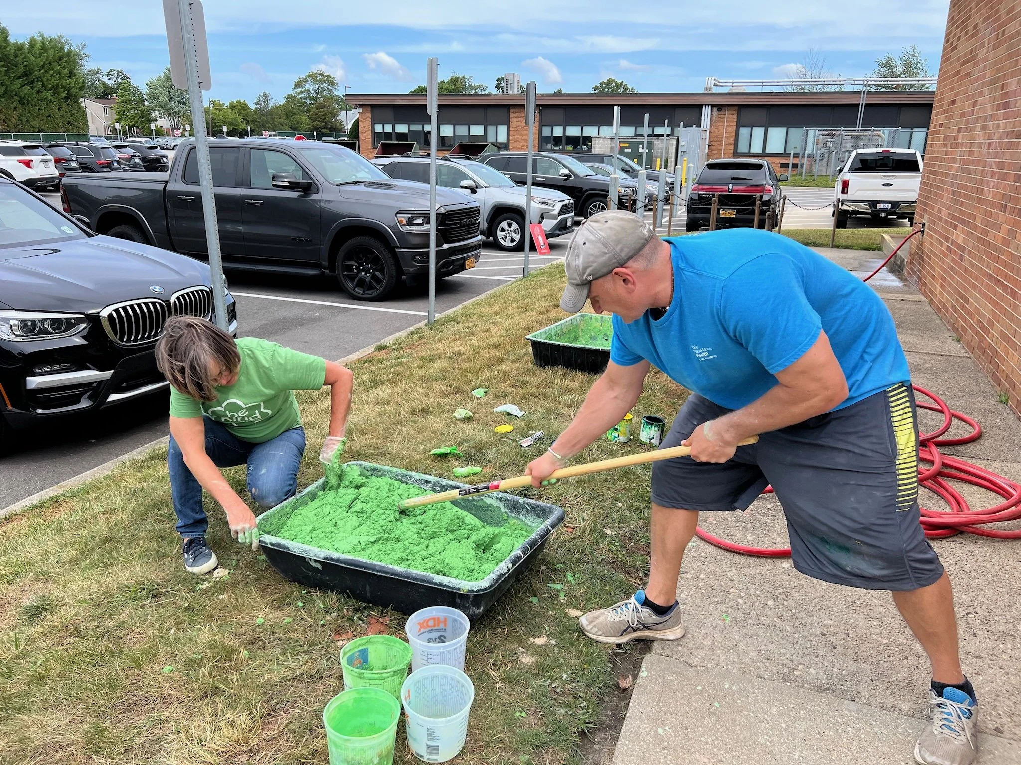 Two people working together on a large green foam project outdoors next to a parking lot, using a long brush and wearing casual clothes.