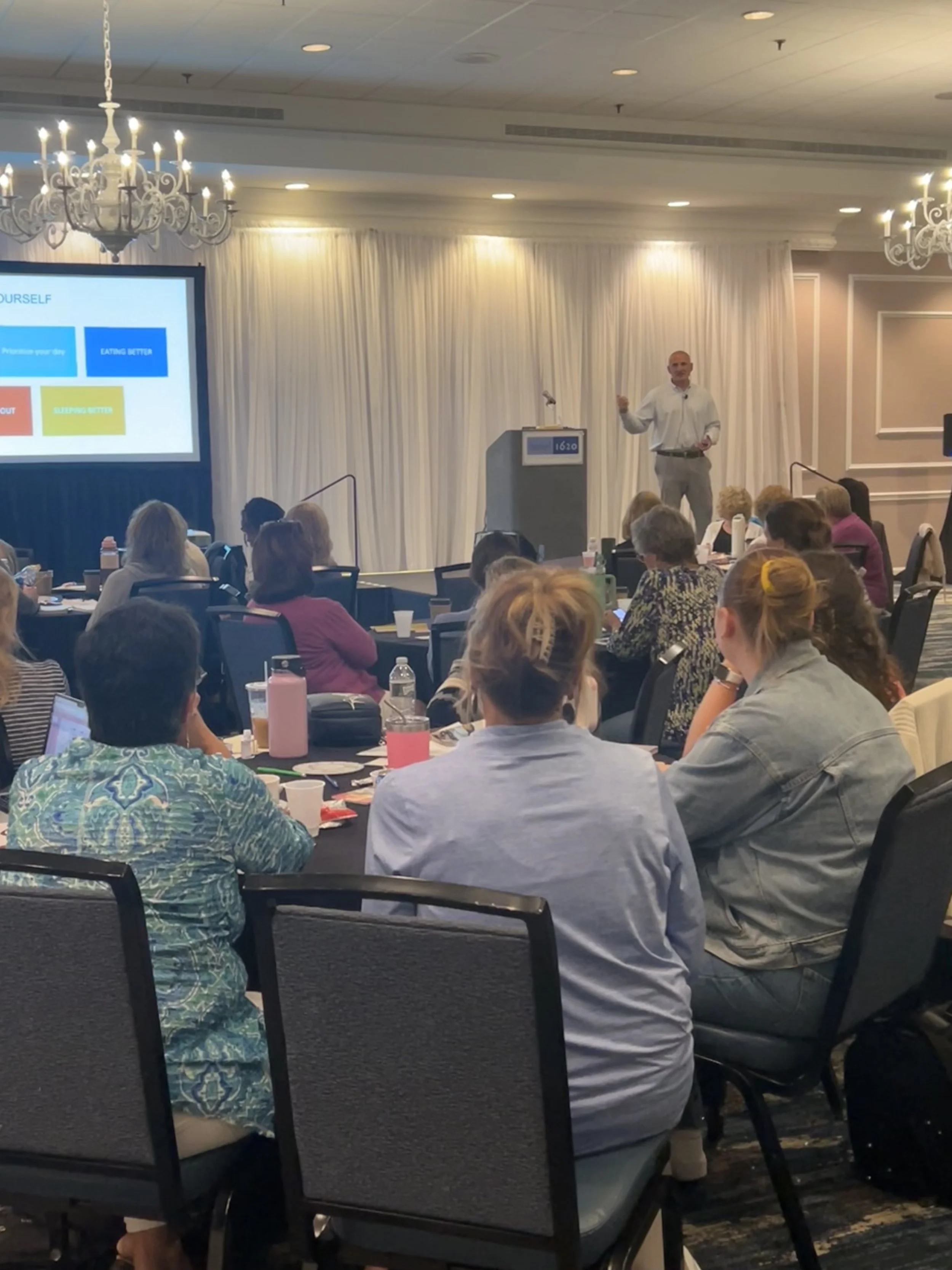 A man giving a presentation to an audience in a conference room with a large screen displaying a slide, and attendees seated at round tables.