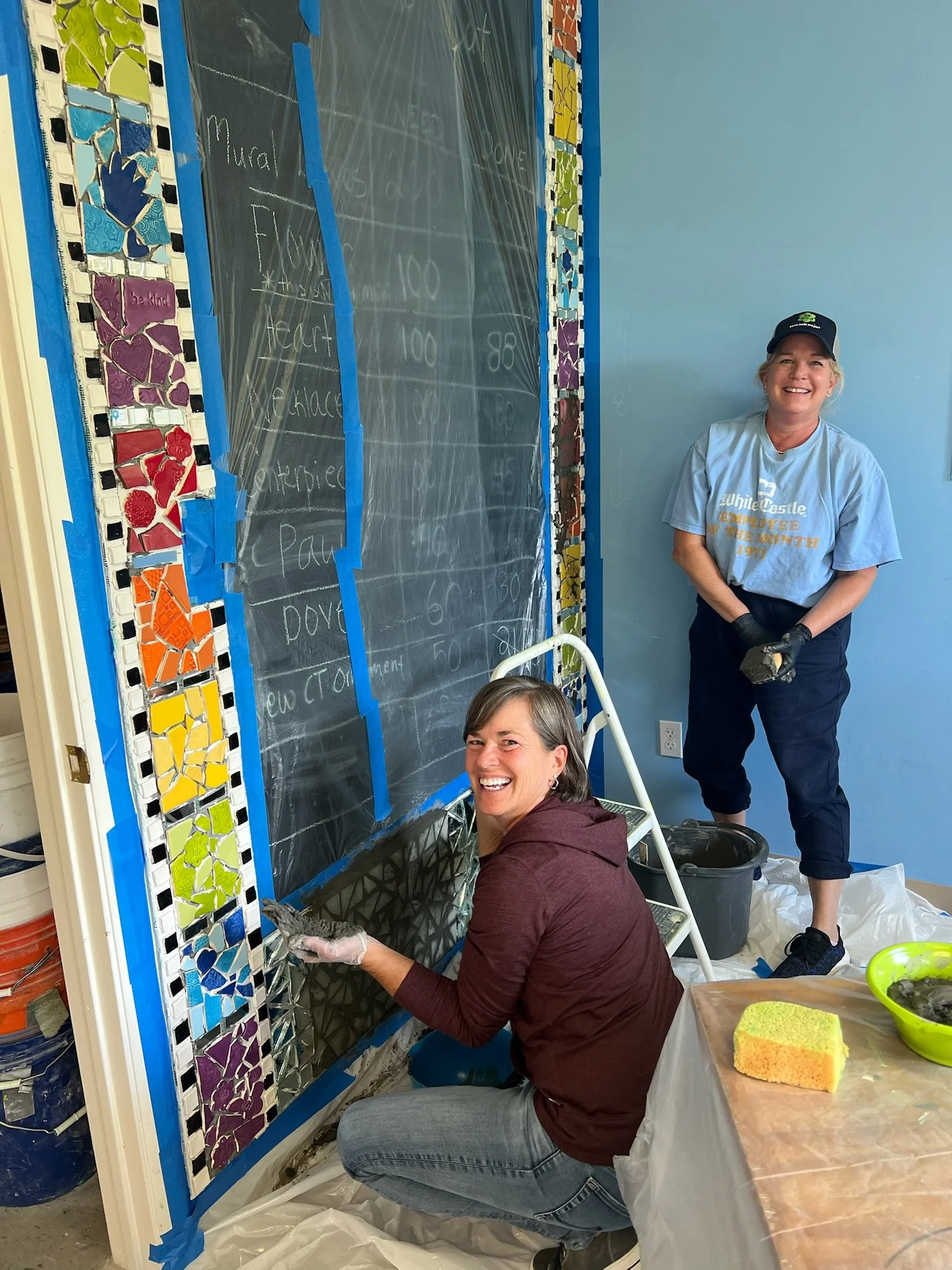 Two women working on a mosaic wall; one is kneeling with a tile in hand, and the other is standing smiling, holding a tool, next to a chalkboard with handwritten notes.