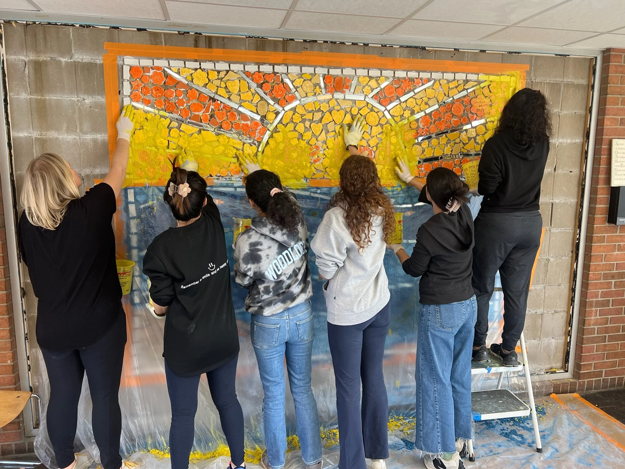 Group of six people painting a large mosaic mural on a wall, using yellow, orange, and blue paint.