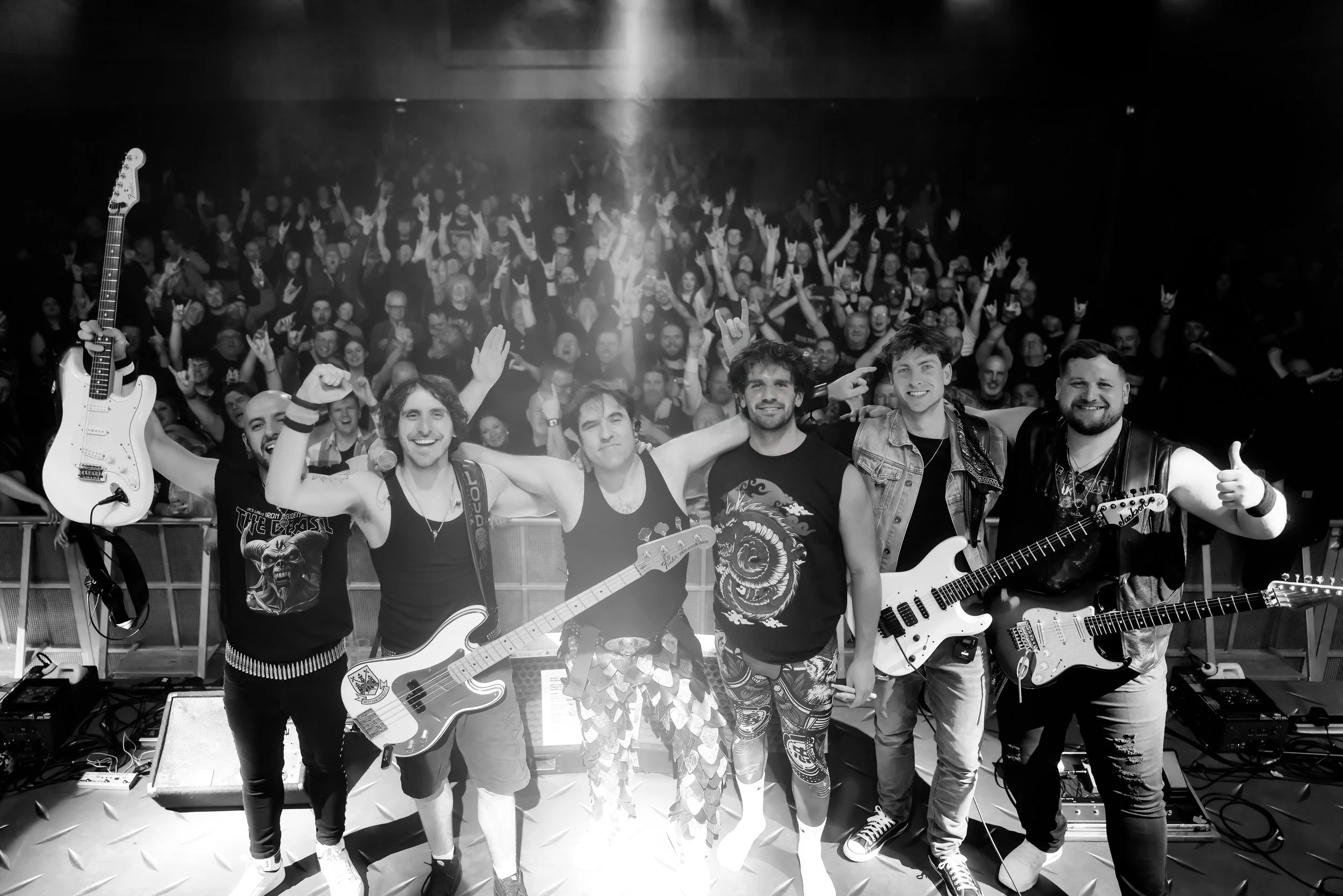 Band of six musicians on stage with guitars, smiling and posing for a photo, with an audience in the background during a concert.