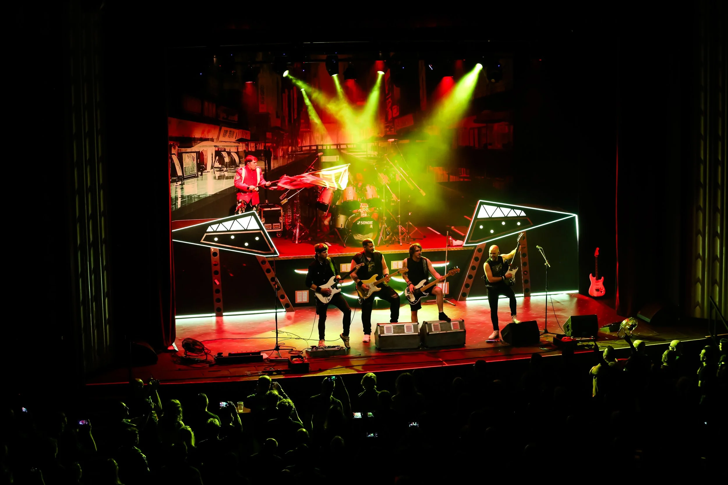 A band performs on stage with colorful lighting, four guitarists at the front and a drummer at the back, in front of an audience in a theater or concert hall.