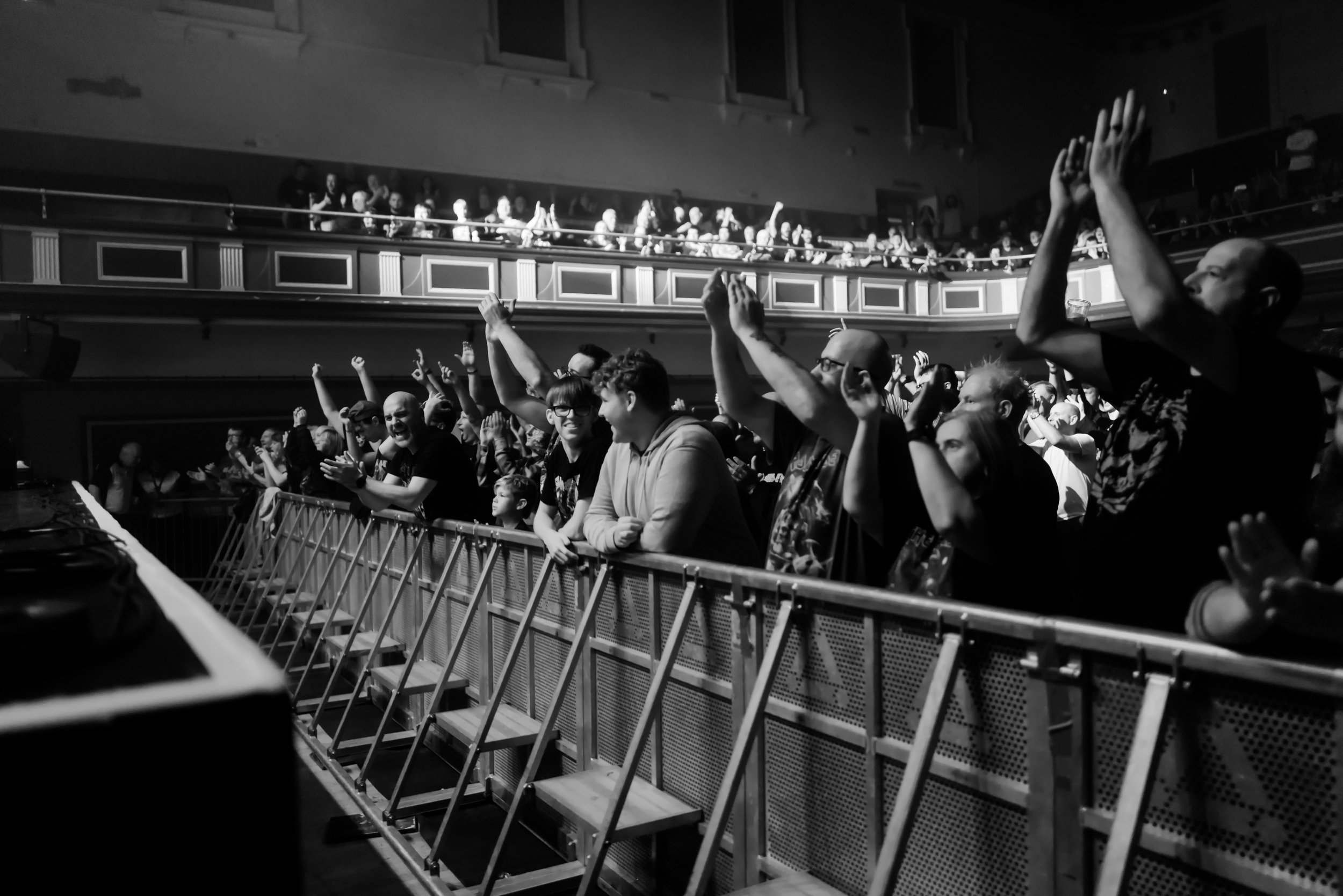 Black and white photo of a crowd at a concert, clapping and cheering with people in the audience and balcony in the background.