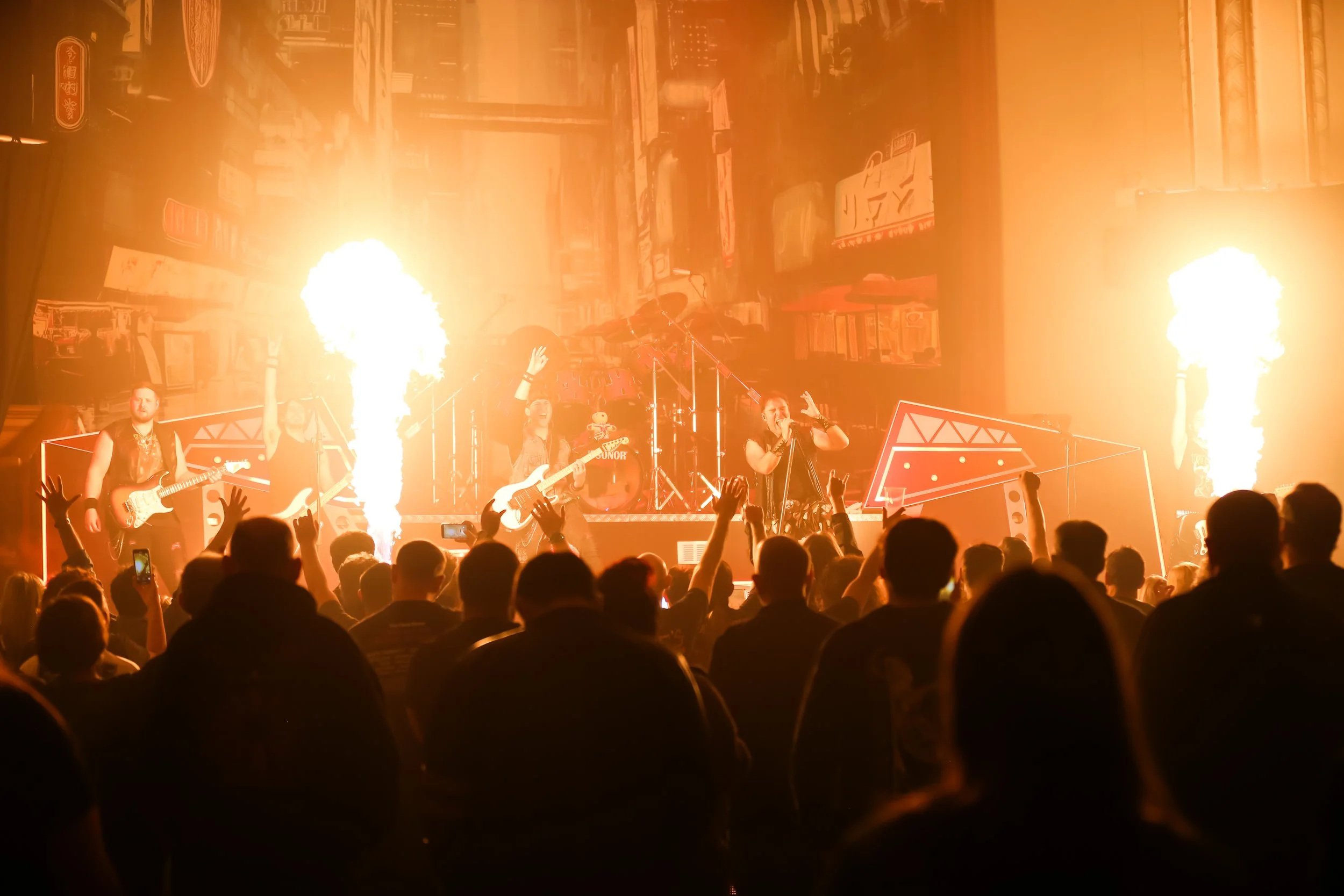A live concert with five band members on stage, surrounded by flames and bright lights. The audience is engaged, with some raising their hands and taking photos.