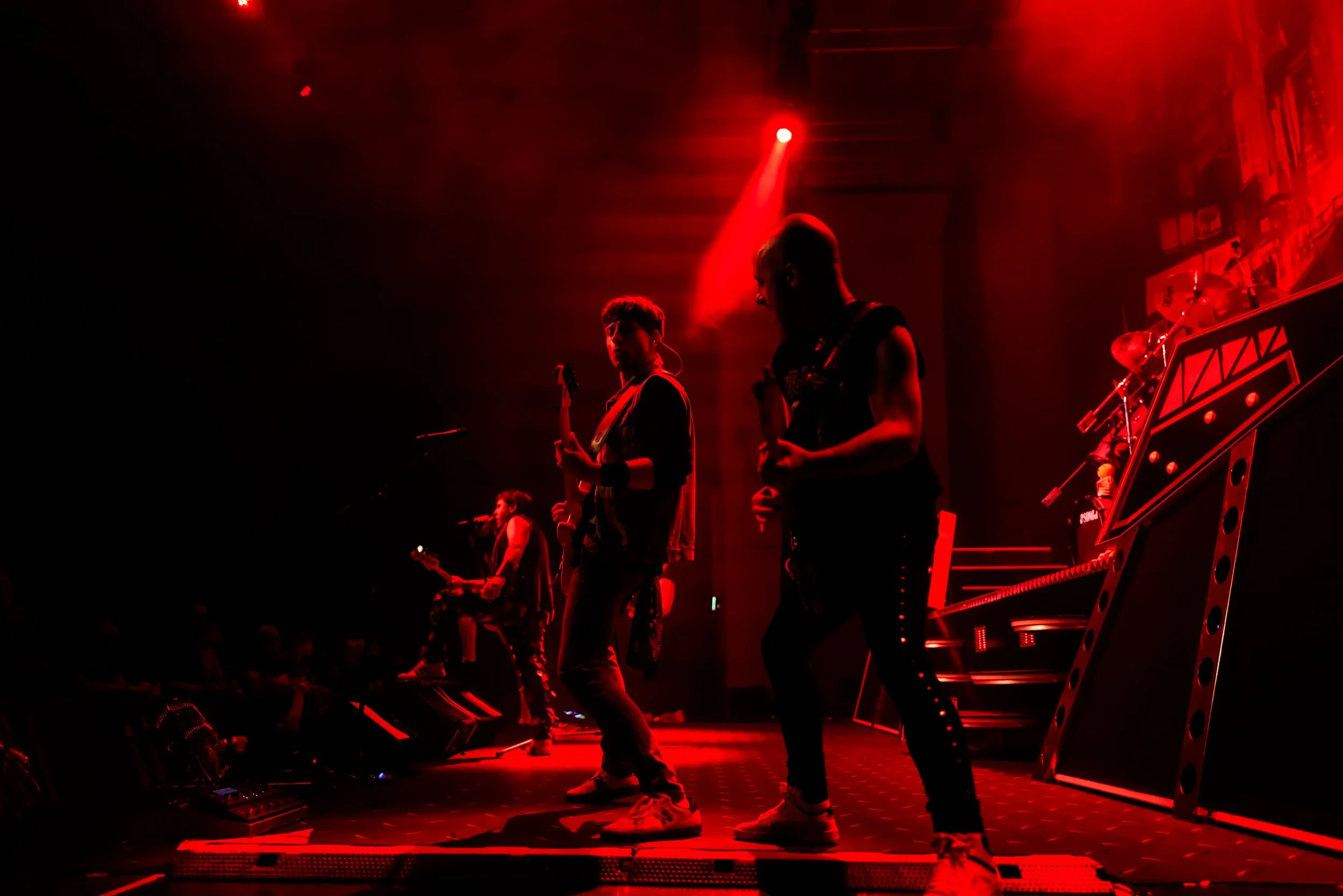 Band performing on stage under red lighting, with musicians playing guitars and drums.