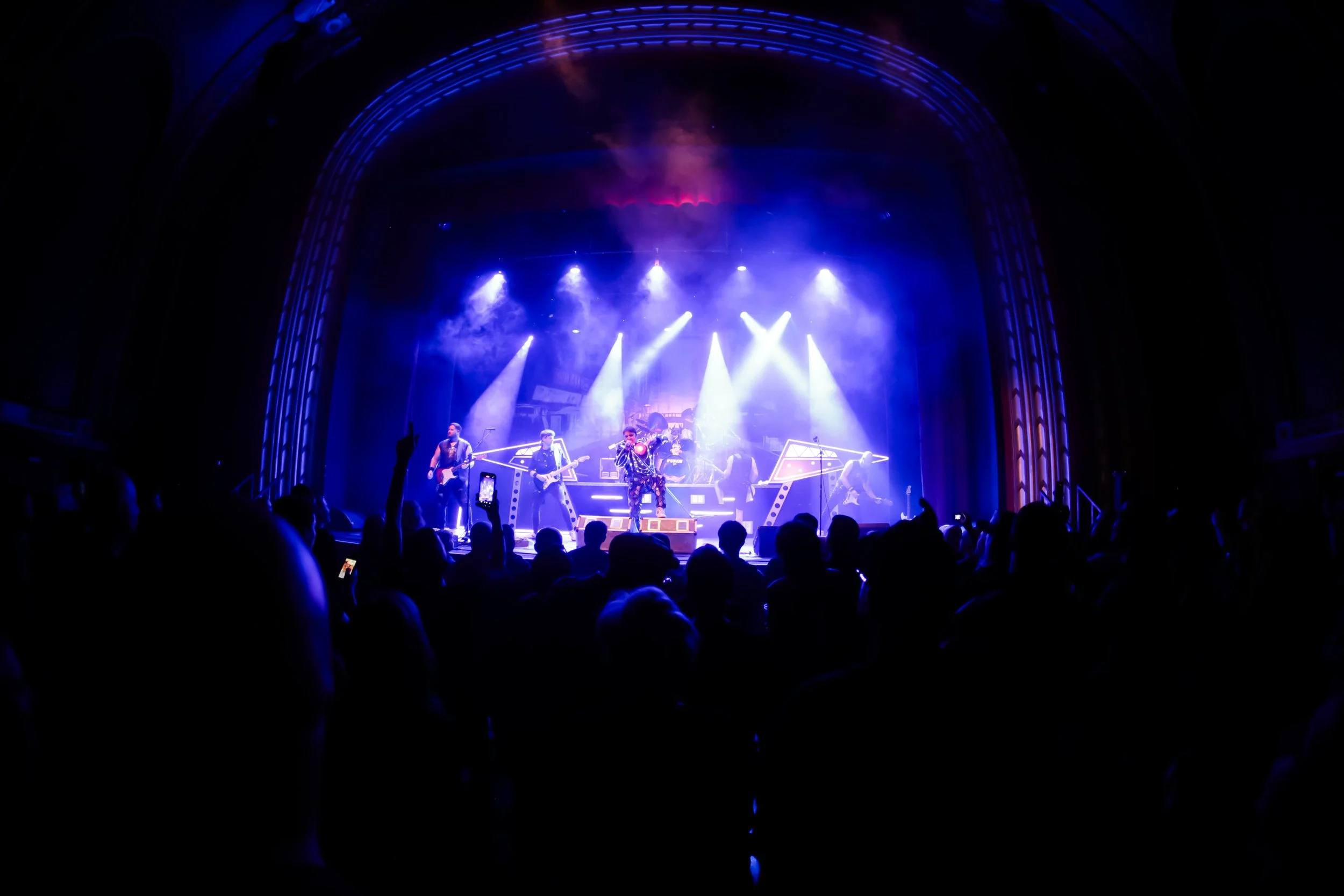 A live concert on stage with band members performing under blue and white stage lights, audience in foreground.