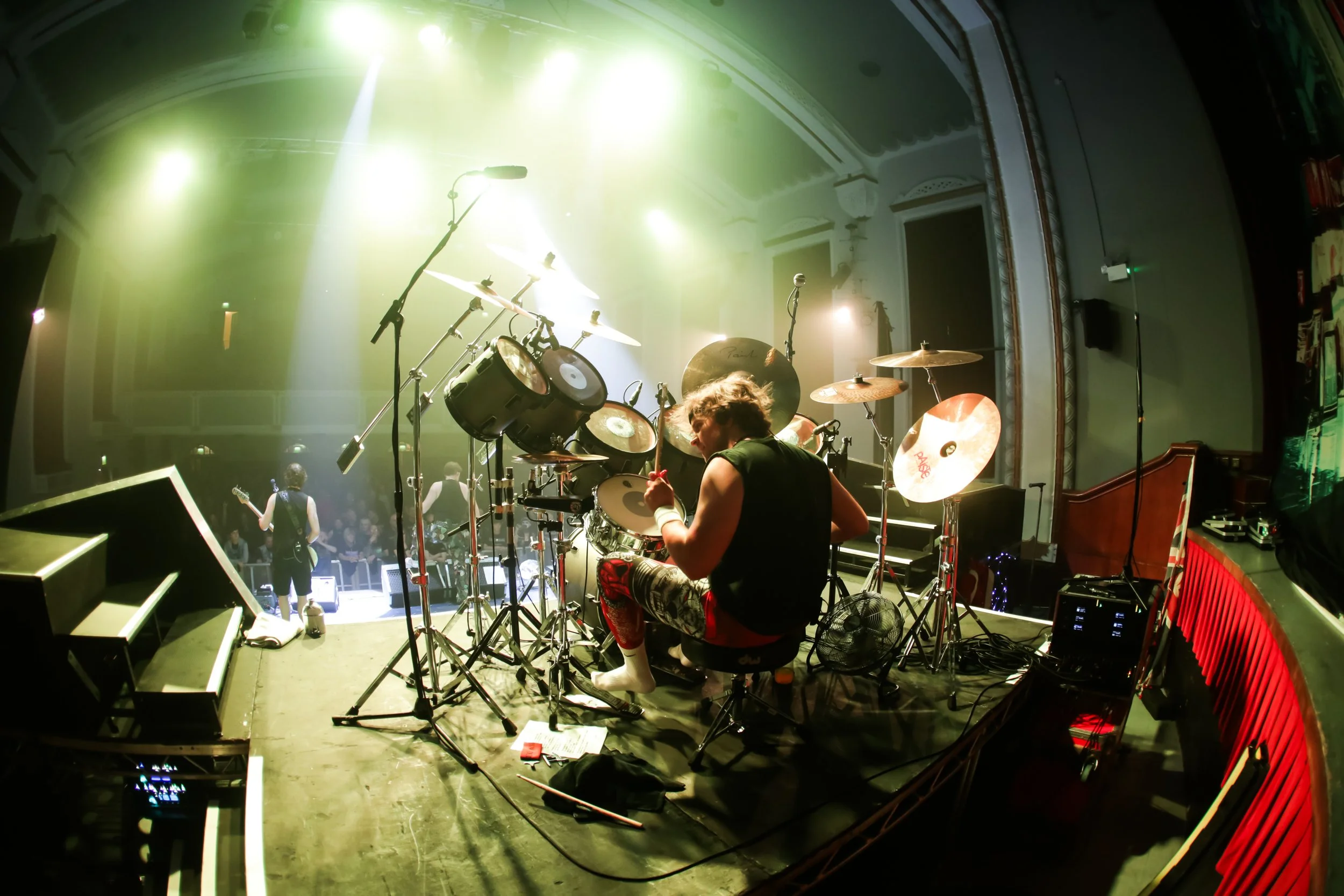 A drummer playing on stage with drums, cymbals, and microphones, illuminated by colorful stage lights before a live audience.