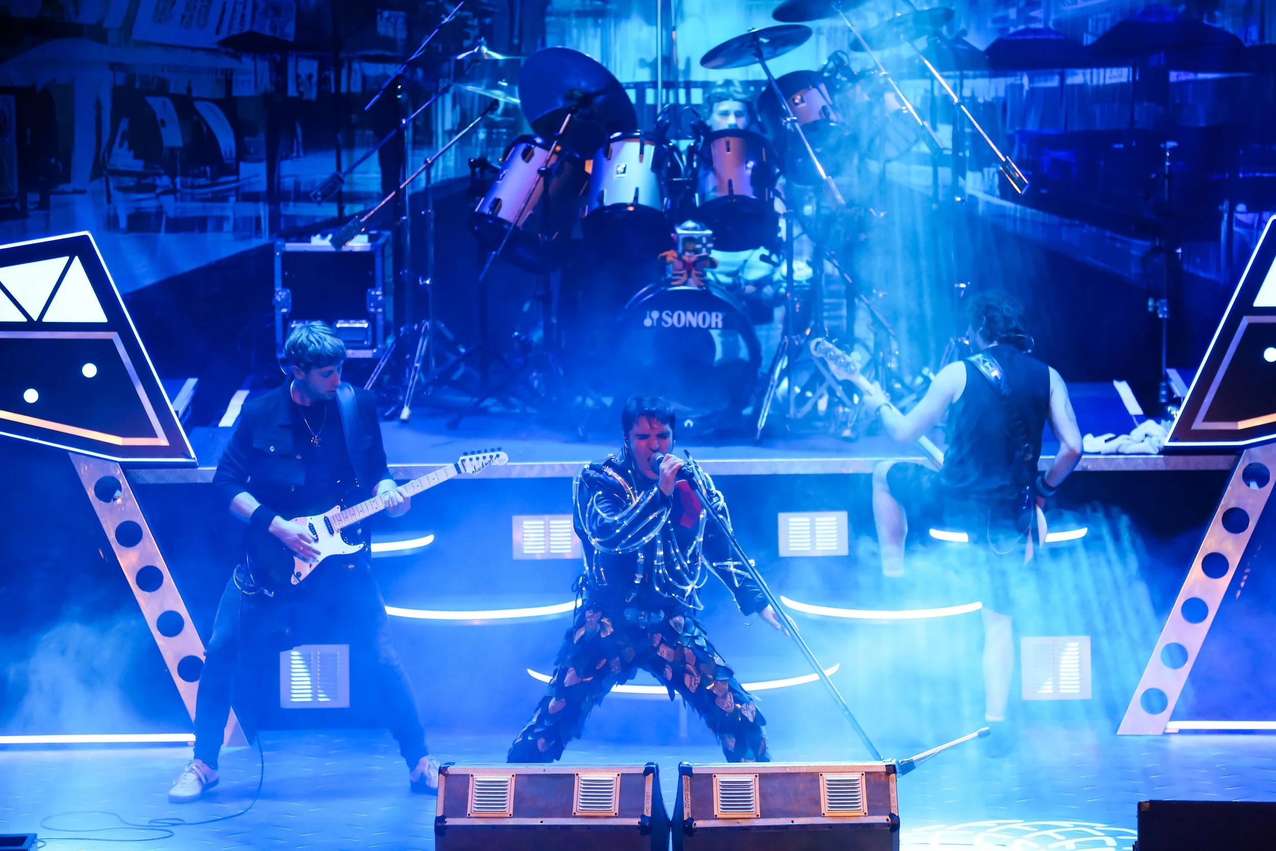 Band performing on stage with a vocalist in the center, guitarist on the left, and bassist on the right, with drums in the background, illuminated by blue stage lighting and fog effects.