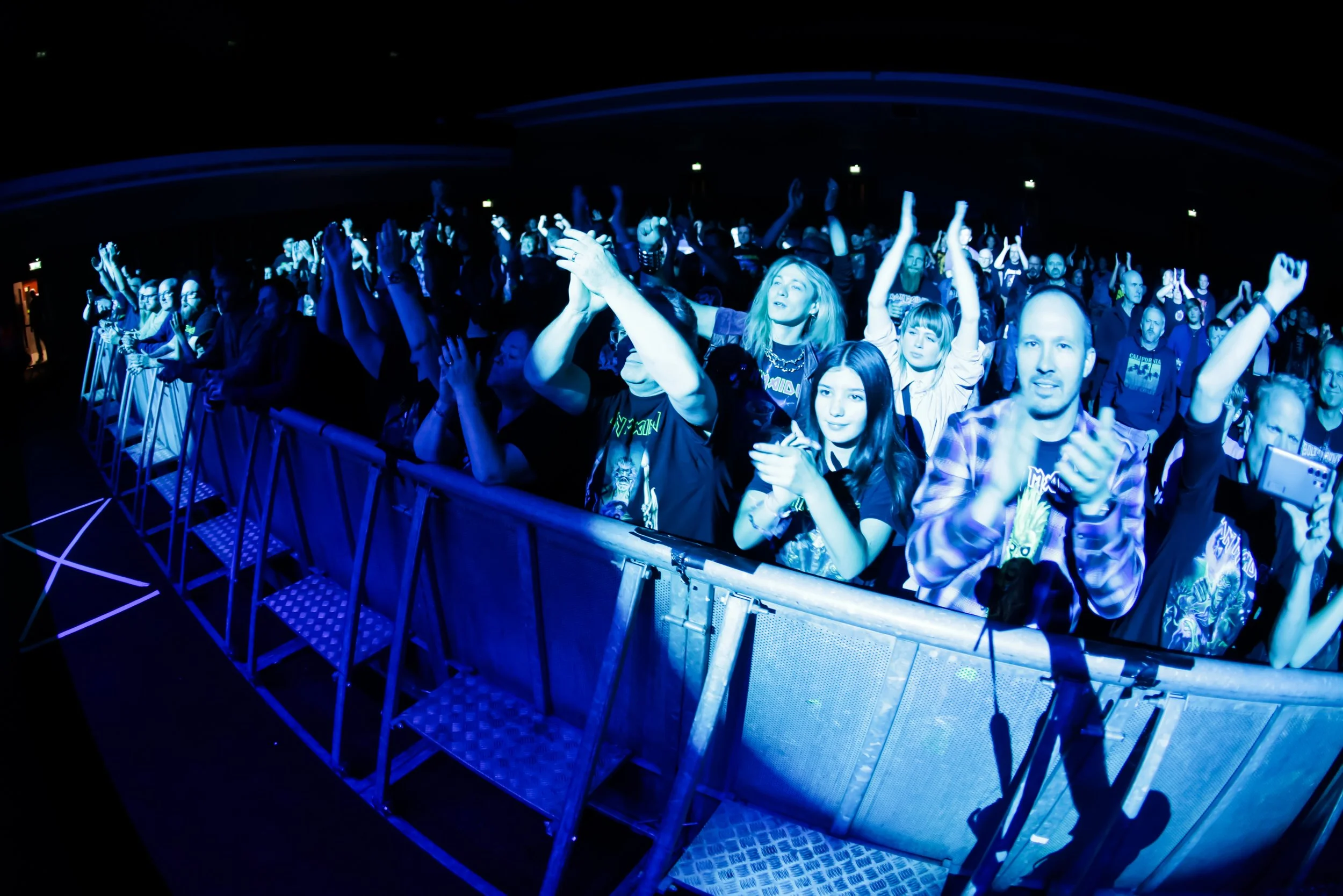 Audience at a concert or event, clapping and cheering near a metal barricade, with blue lighting highlighting their faces.