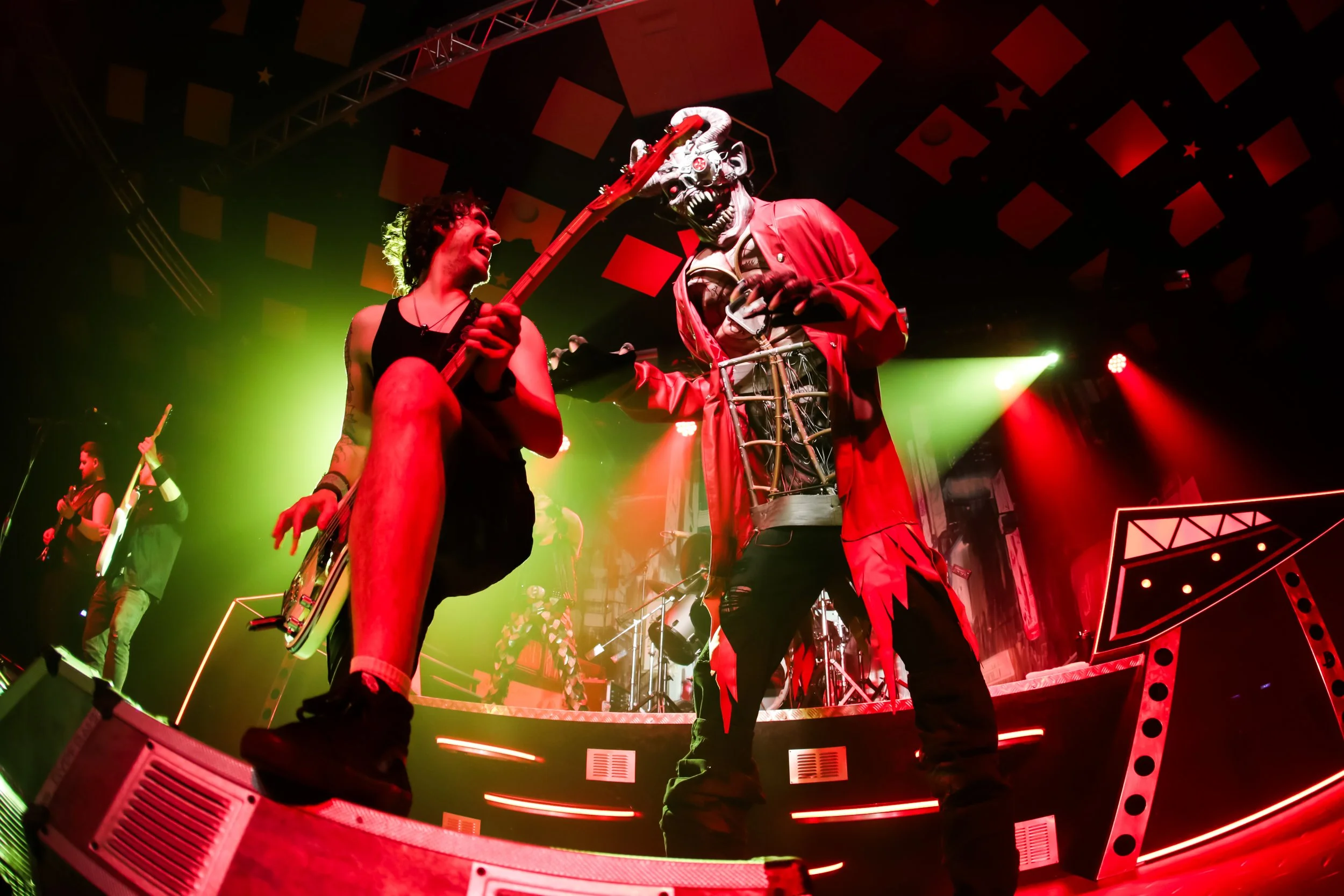 A live rock concert with performers on stage, including a person in a demon mask and red clothing, and another person with a guitar. The stage is lit with vibrant red and green lights.
