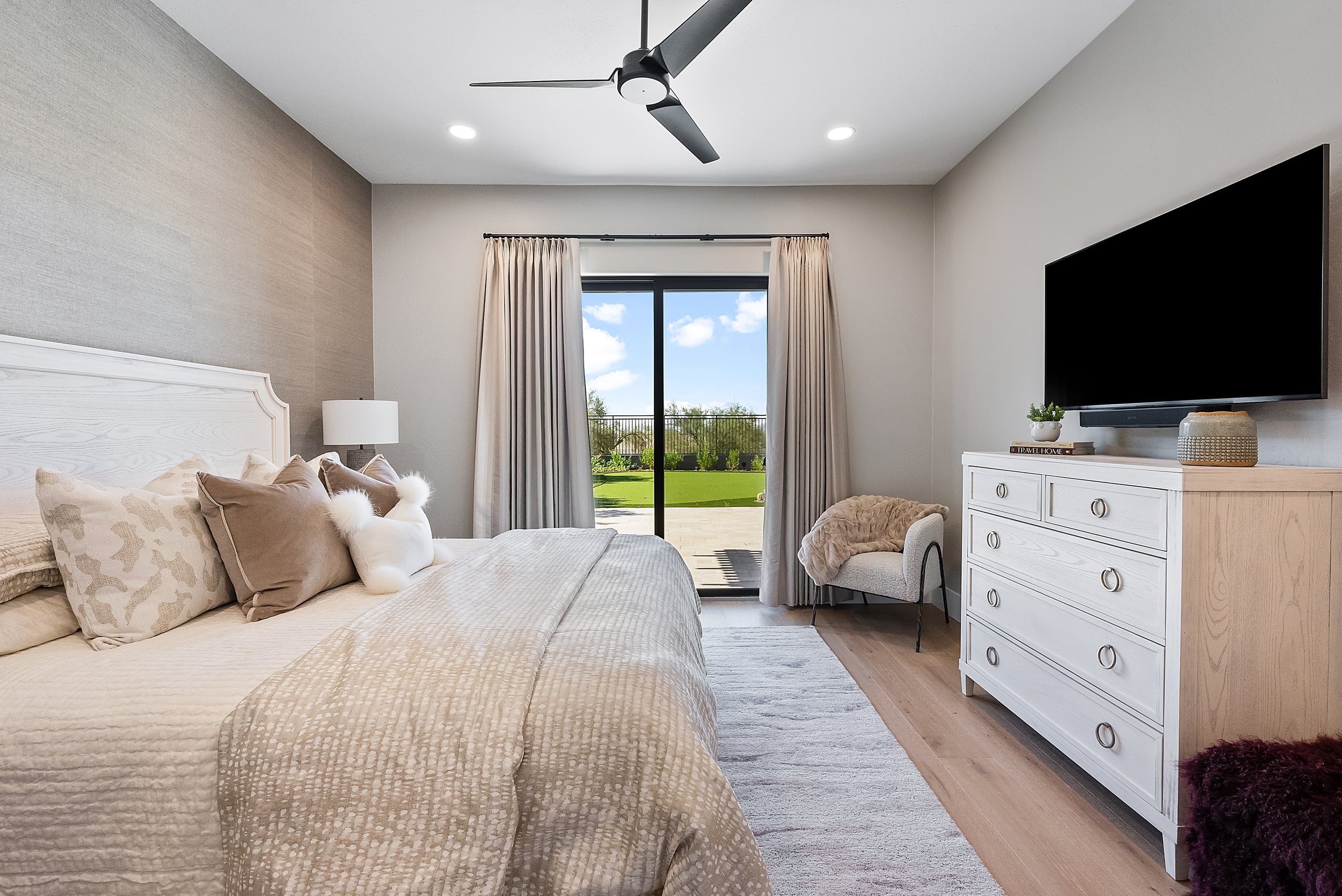 A bedroom with a bed, pillows, and a blanket, a dresser with a small plant and books, a large flat-screen TV mounted on the wall, beige curtains, a sliding glass door showing an outdoor view of green grass, trees, and a partly cloudy sky, a ceiling f