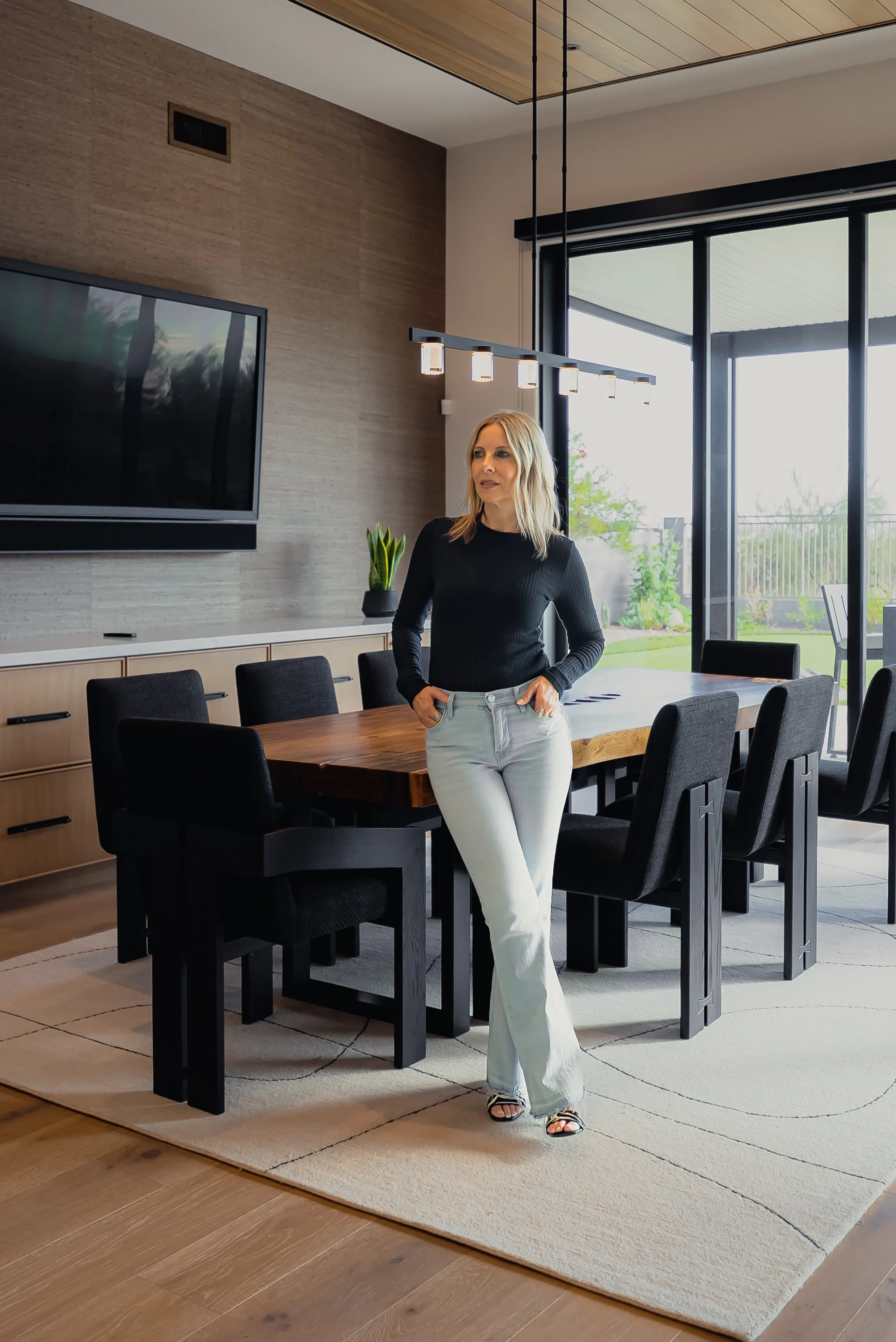 A woman with blonde hair, wearing a black long-sleeve top and white pants, standing in a modern dining room with black chairs, a wooden table, and a large window with sliding doors.