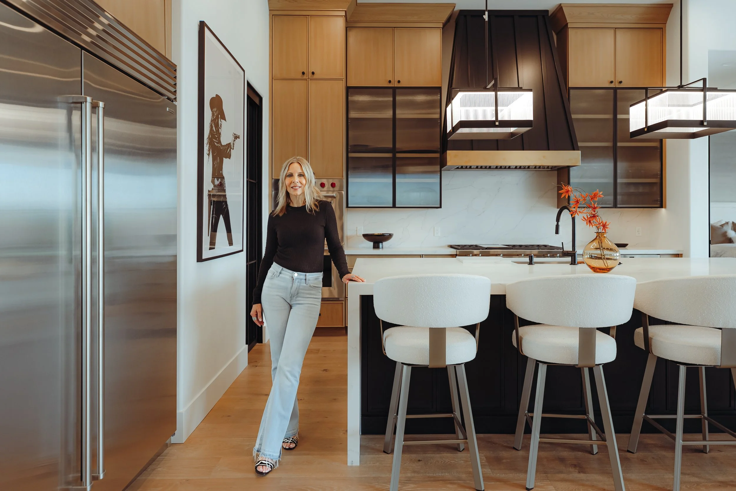 Woman in black sweater and white pants standing in modern kitchen with wooden cabinets, white marble backsplash, black accents, and a vase with orange flowers on the counter.