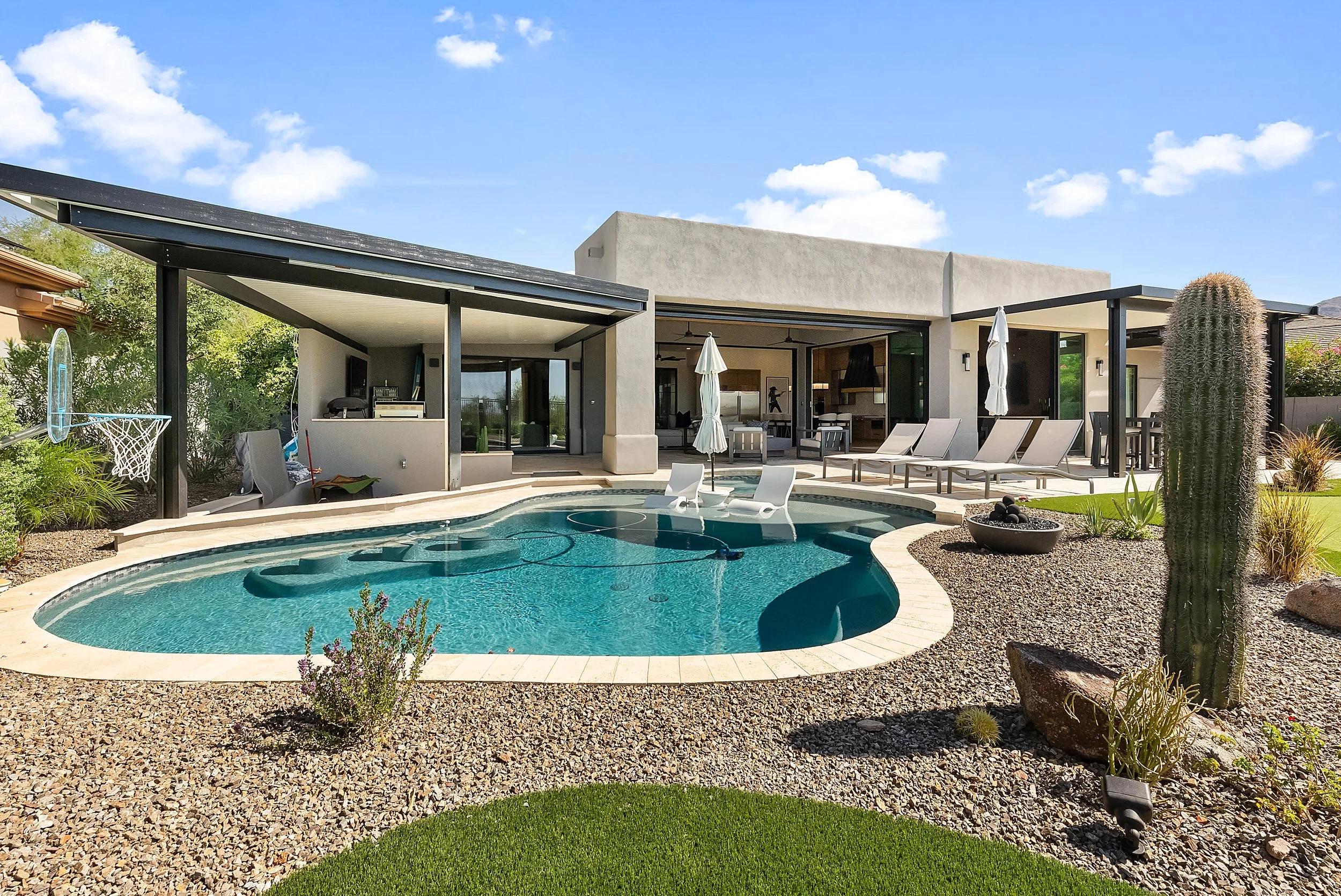 Modern backyard with a swimming pool, surrounded by patio furniture, cactus plants, and a covered outdoor living area with chairs and umbrellas, under a blue sky with scattered clouds.