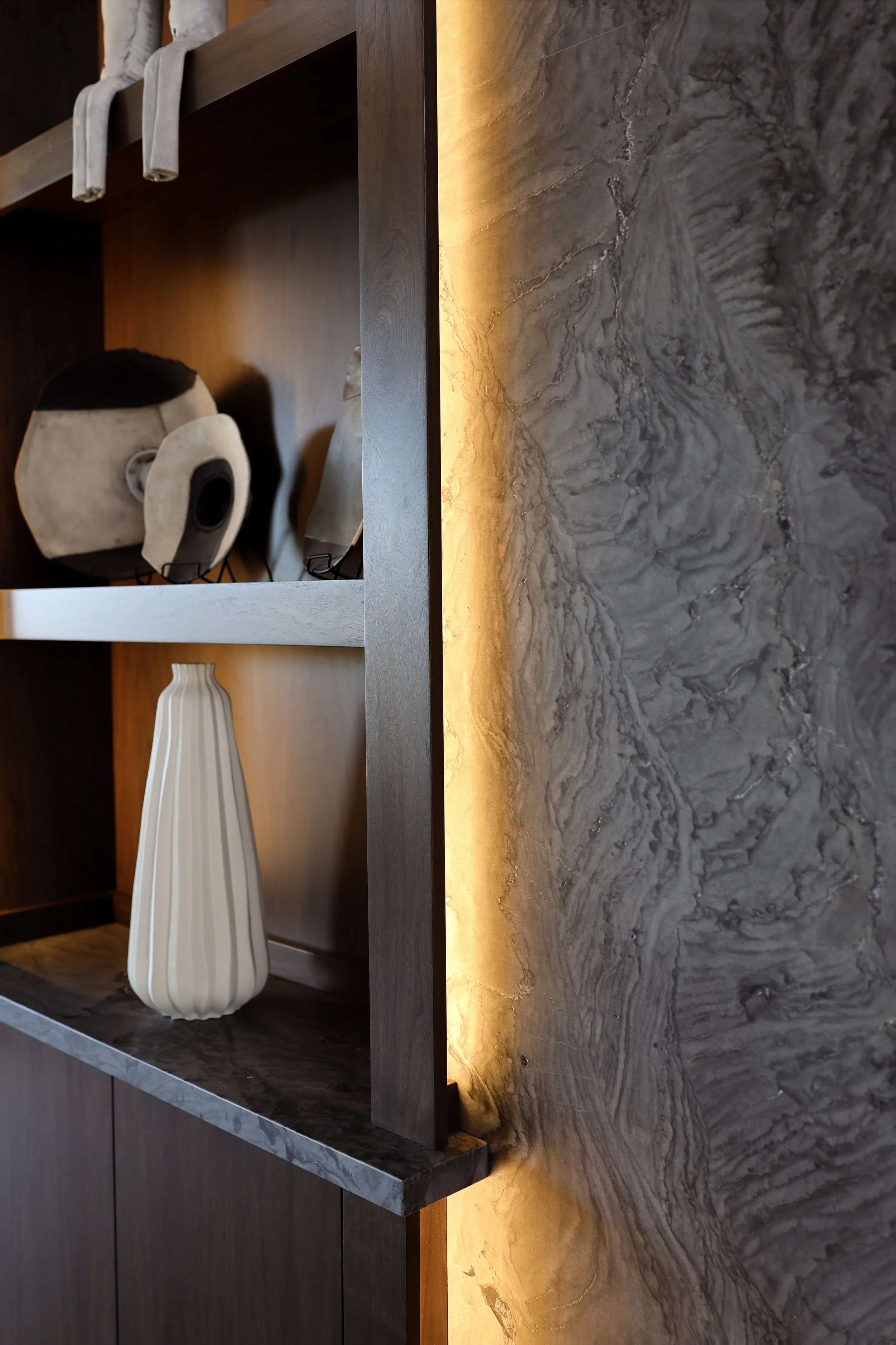 A close-up view of a modern interior shelf decorated with vases and sculptures, with warm lighting accentuating a textured stone wall behind it.