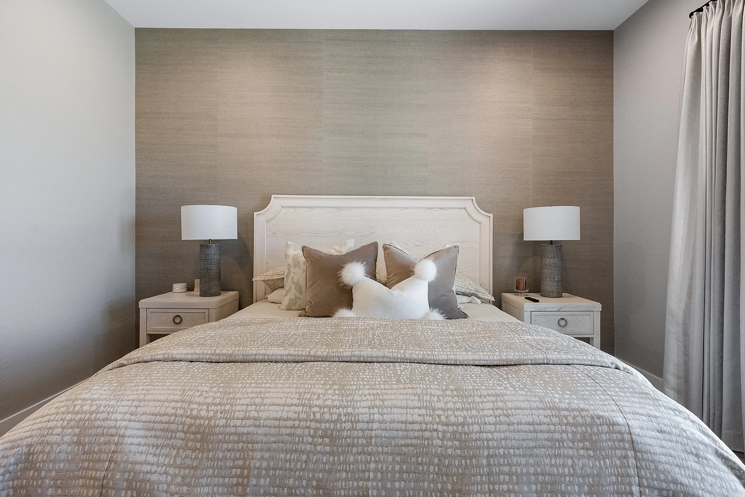 A neatly made bed with beige and cream linens, multiple decorative pillows including fur and patterned ones, flanked by two white nightstands with lamps on each side, against a neutral wall with a beige painted section, and cream curtains on the righ