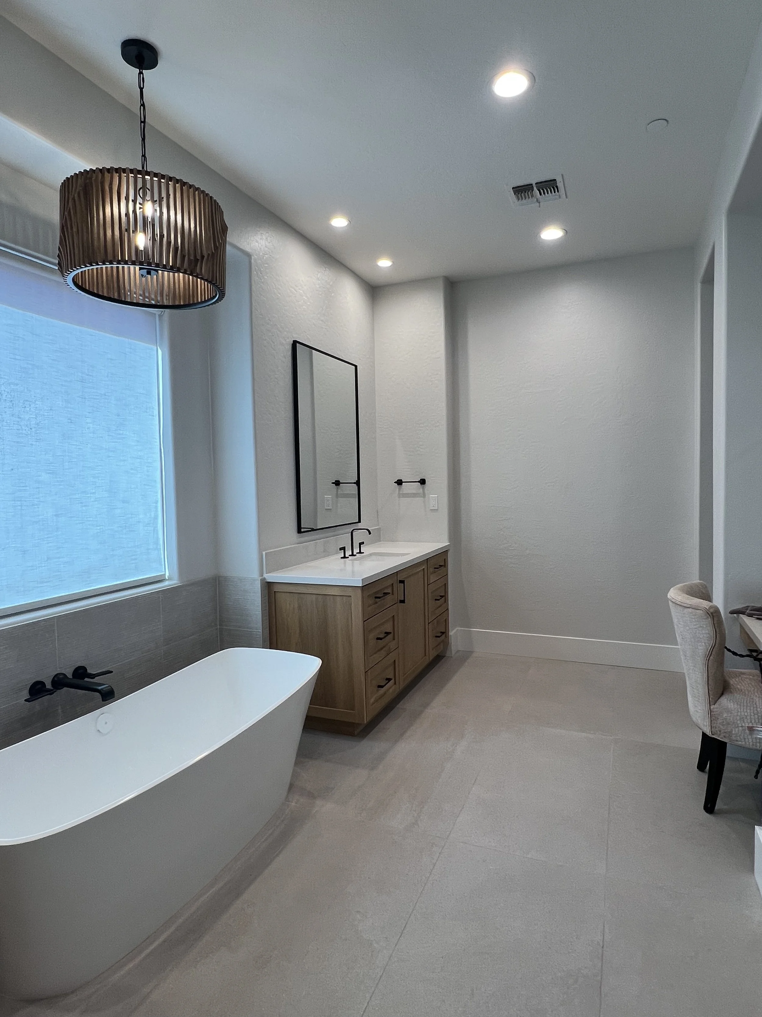 Modern bathroom with a white freestanding bathtub, wooden vanity with a sink, large mirror, black towel bars, and a window with a roller shade. Recessed ceiling lights and a pendant light fixture.