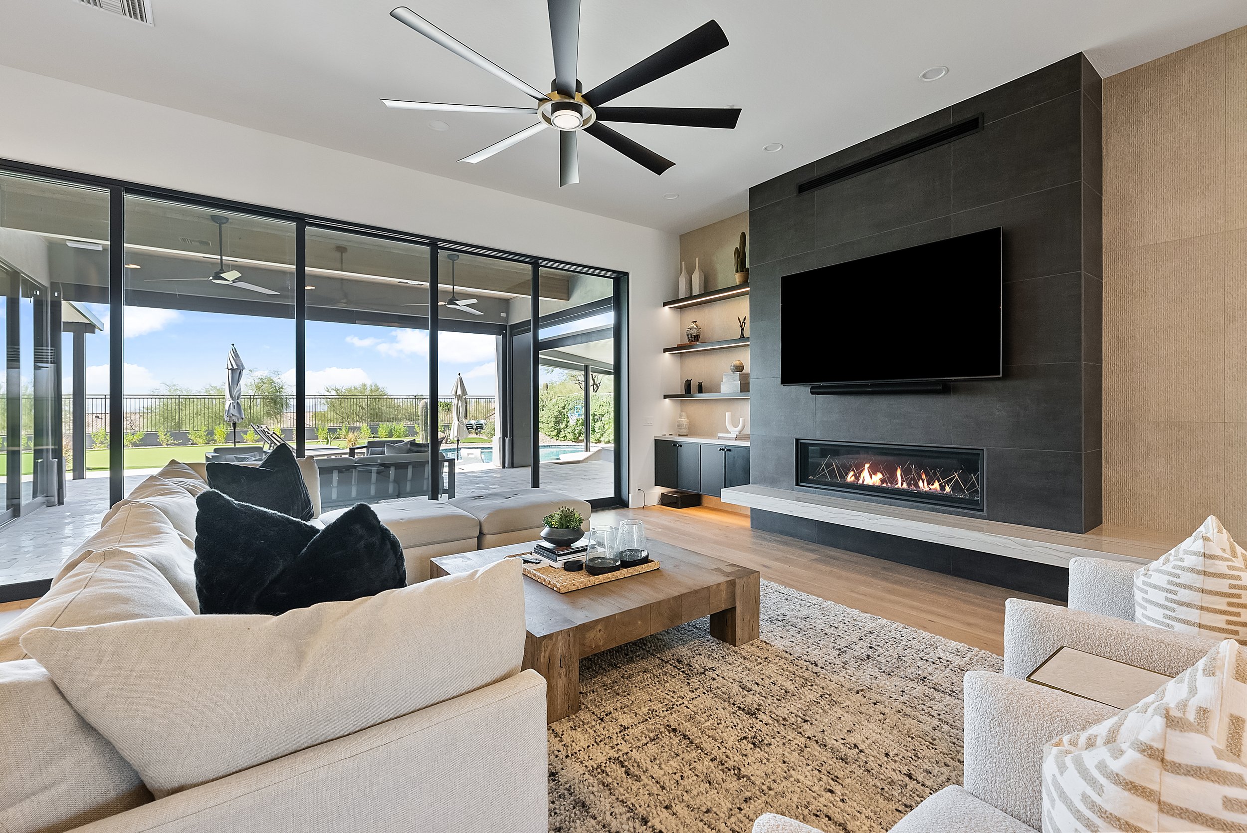 Modern living room with large sectional sofa, black throw pillows, wooden coffee table with decorative items, fireplace, wall-mounted TV, glass sliding doors with outdoor patio and pool, ceiling fan, shelves with decorative objects, and large windows