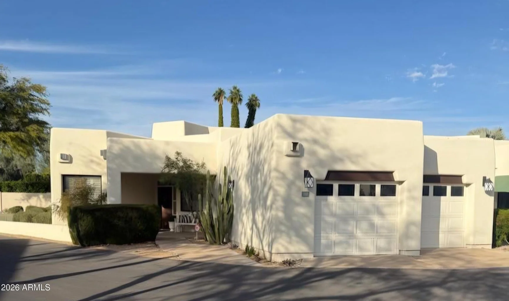 A modern, minimalist house with a white stucco exterior, flat rooflines, and small rectangular windows. The house has a garage door and a front porch with a white bench. Tall cacti and palm trees are visible on the property, with shadows cast on the 