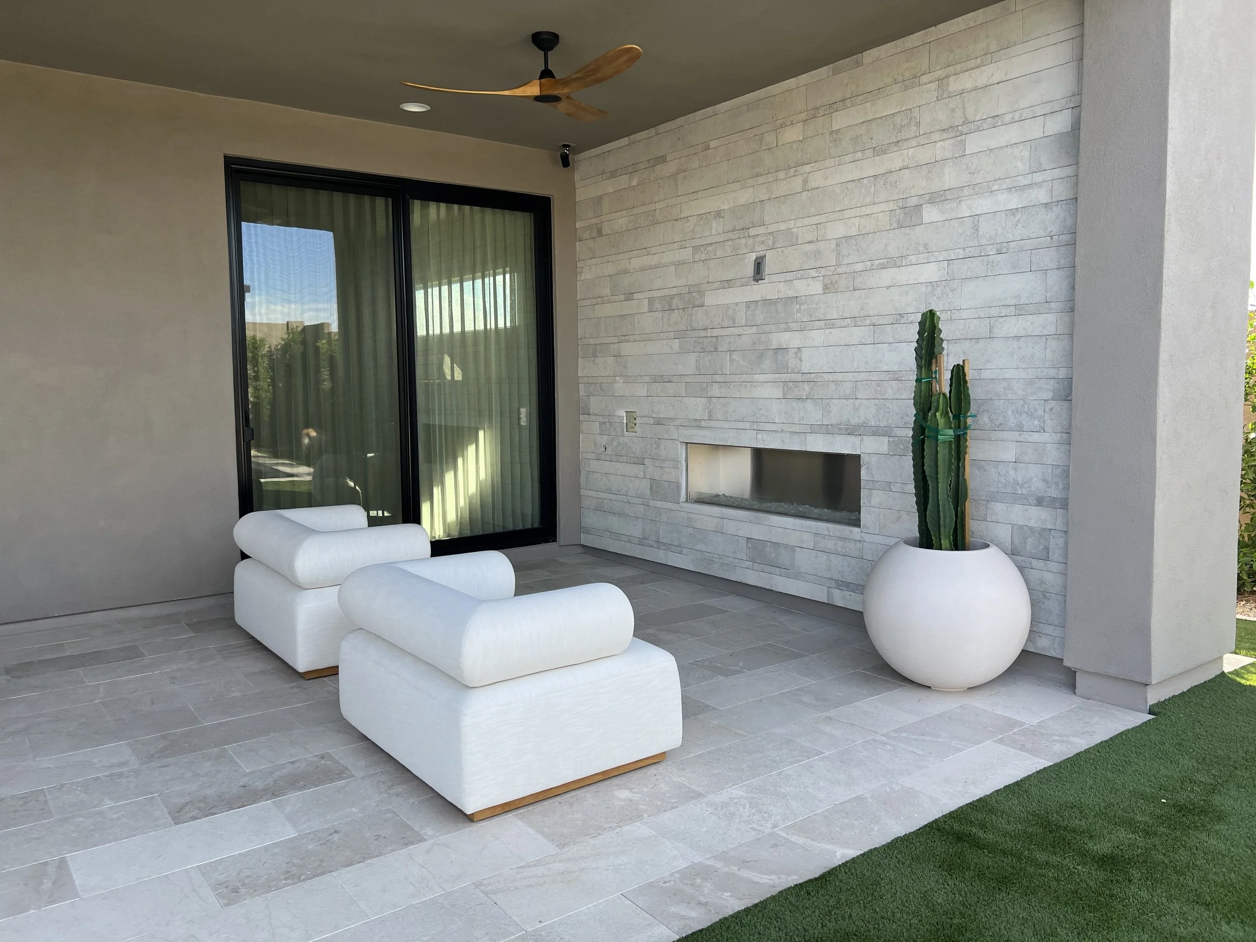 A modern backyard patio featuring a white sectional lounge chair, a large round white planter with a tall cactus, and a stone wall with a rectangular opening. There is a ceiling fan with wooden blades and sliding glass doors leading inside.