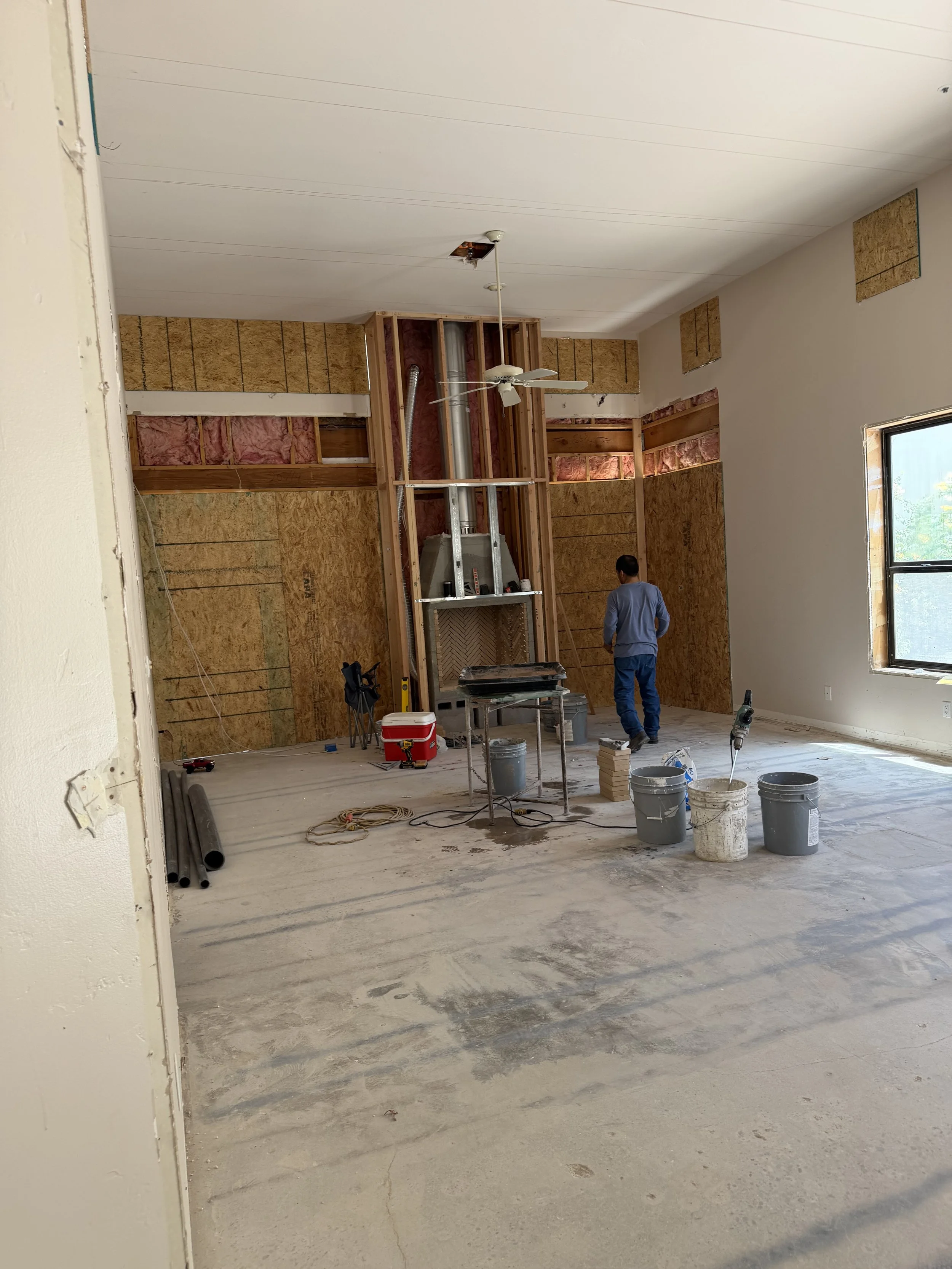 Interior of a room under renovation with exposed framing, a person working, construction tools, buckets, and partially finished walls.