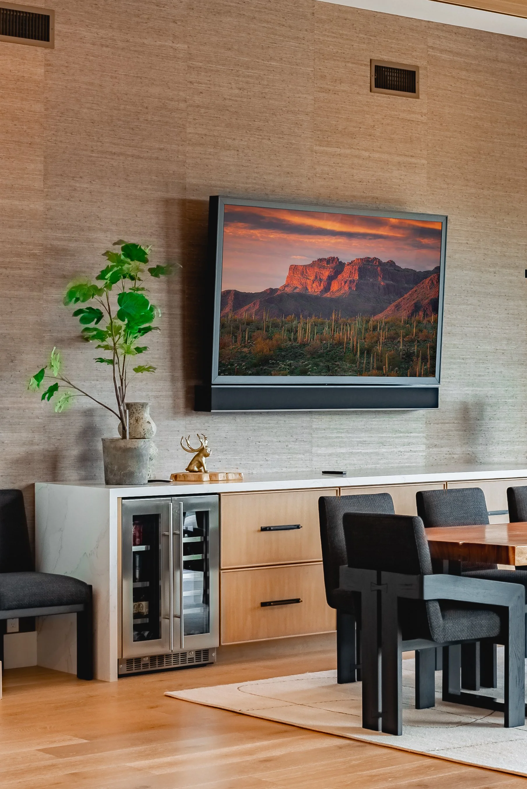 Modern dining room with wooden floors, a large dining table with black chairs, a decorative plant in a pot, and a framed mountain landscape picture on the beige wall.