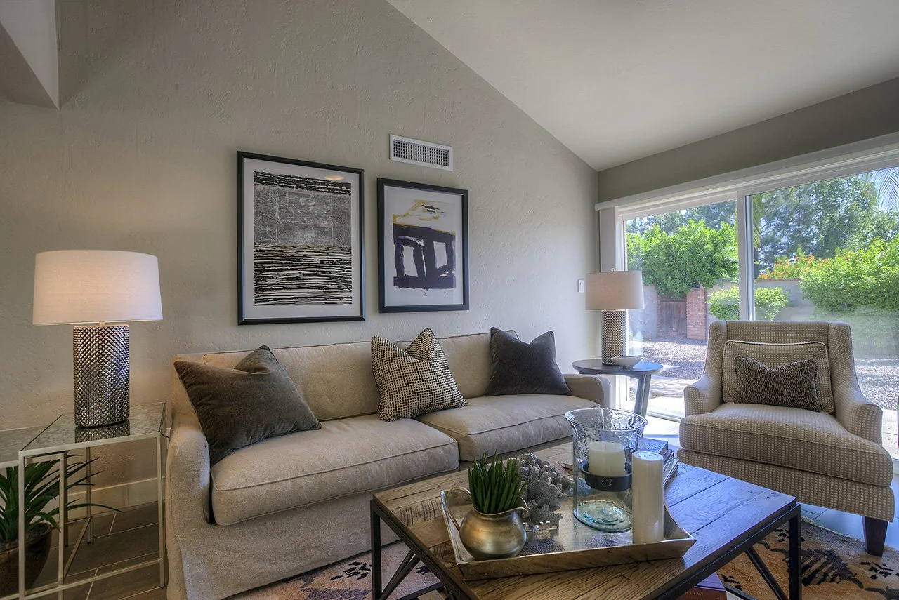 Living room with beige sofa and armchair, abstract artwork on wall, lamps on side tables, coffee table with decorations, large window showing outdoor greenery.