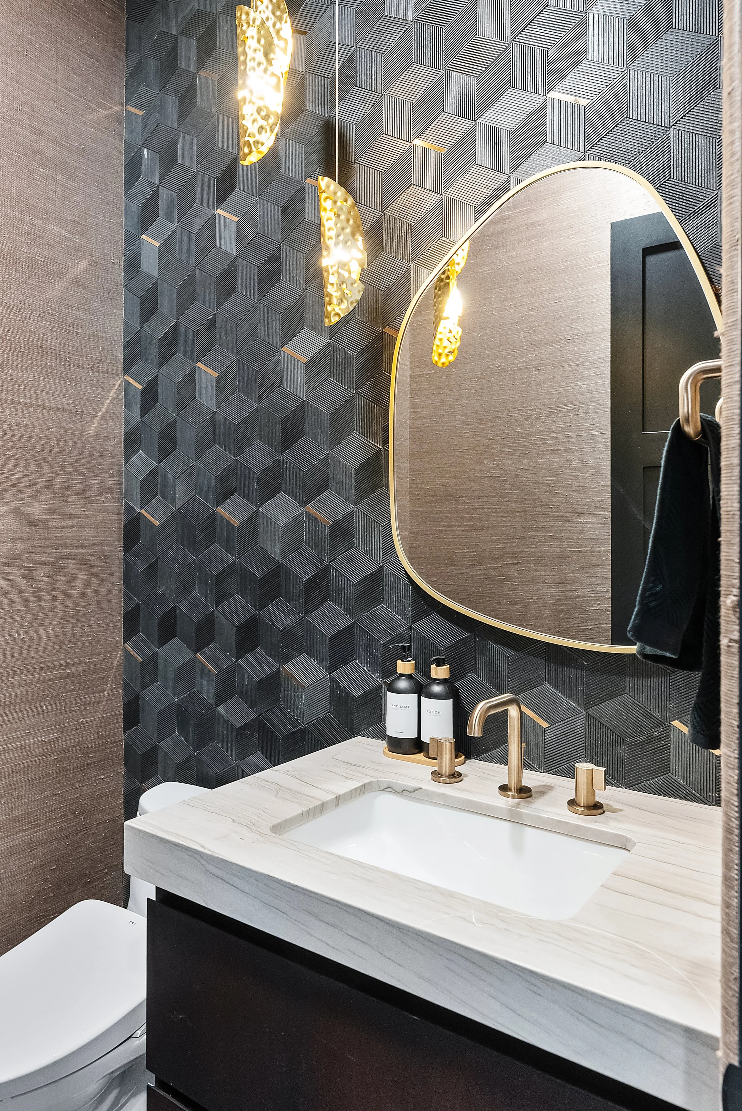 Modern bathroom with black geometric patterned wall, gold fixtures, and a marble countertop with a sink. There are hand soap and lotion bottles on the counter and a mirror above it. A toilet is visible in the lower left corner.