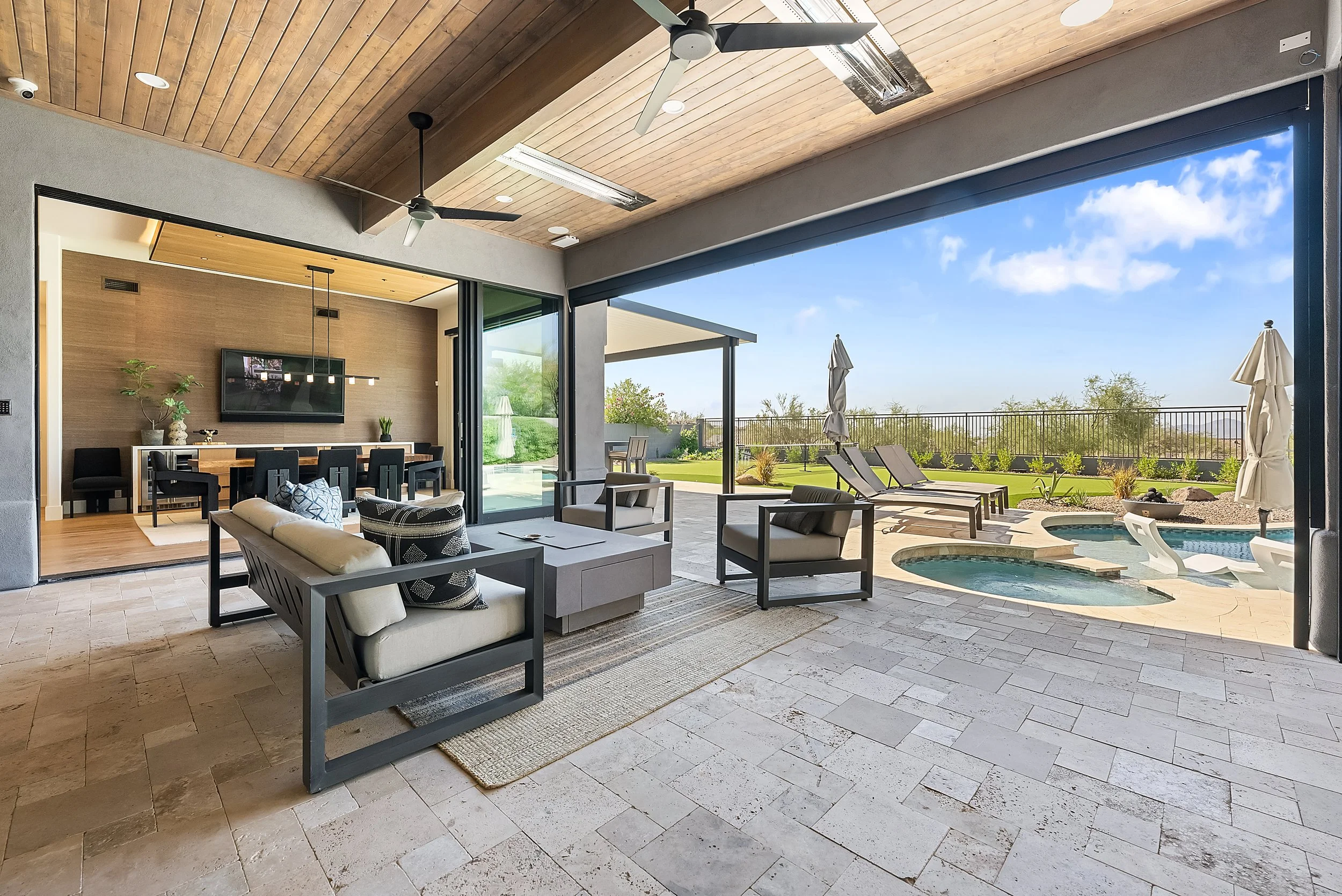 The image shows a spacious outdoor patio area with a swimming pool, lounge chairs, and an open view of a bright blue sky with some clouds and greenery in the background. The patio is connected to an indoor living space through large sliding glass doo