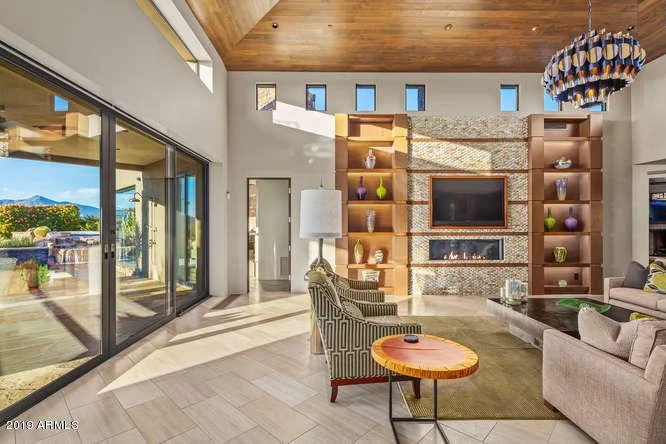 Living room with a high wooden ceiling, large sliding glass door, and a built-in entertainment wall with shelves and decorative vases, furnished with sofas and chairs.