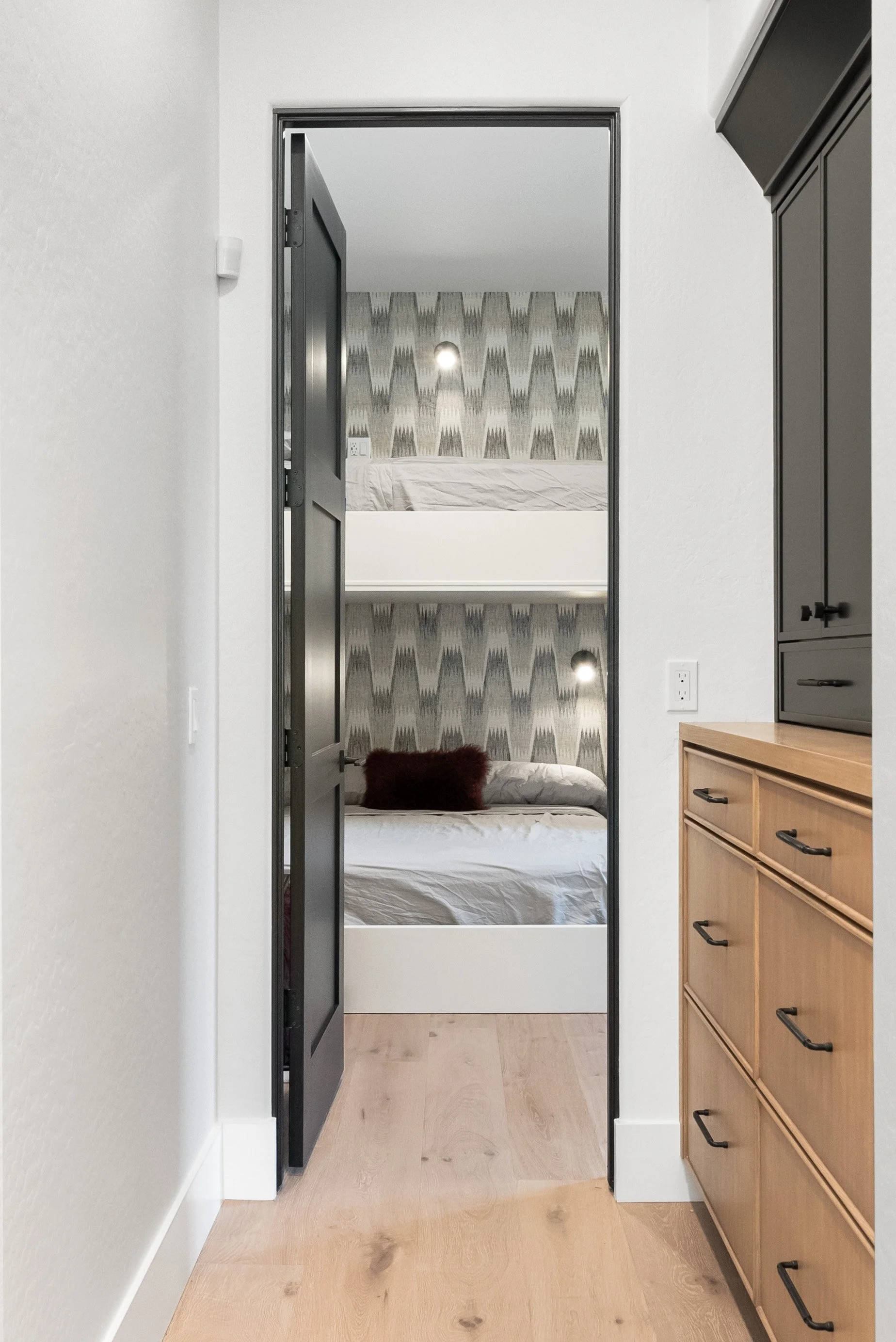 View through an open black door into a bedroom with a bed against a wall with patterned wallpaper, white bedding, a red pillow, and a mirror reflecting part of the room and ceiling lights.