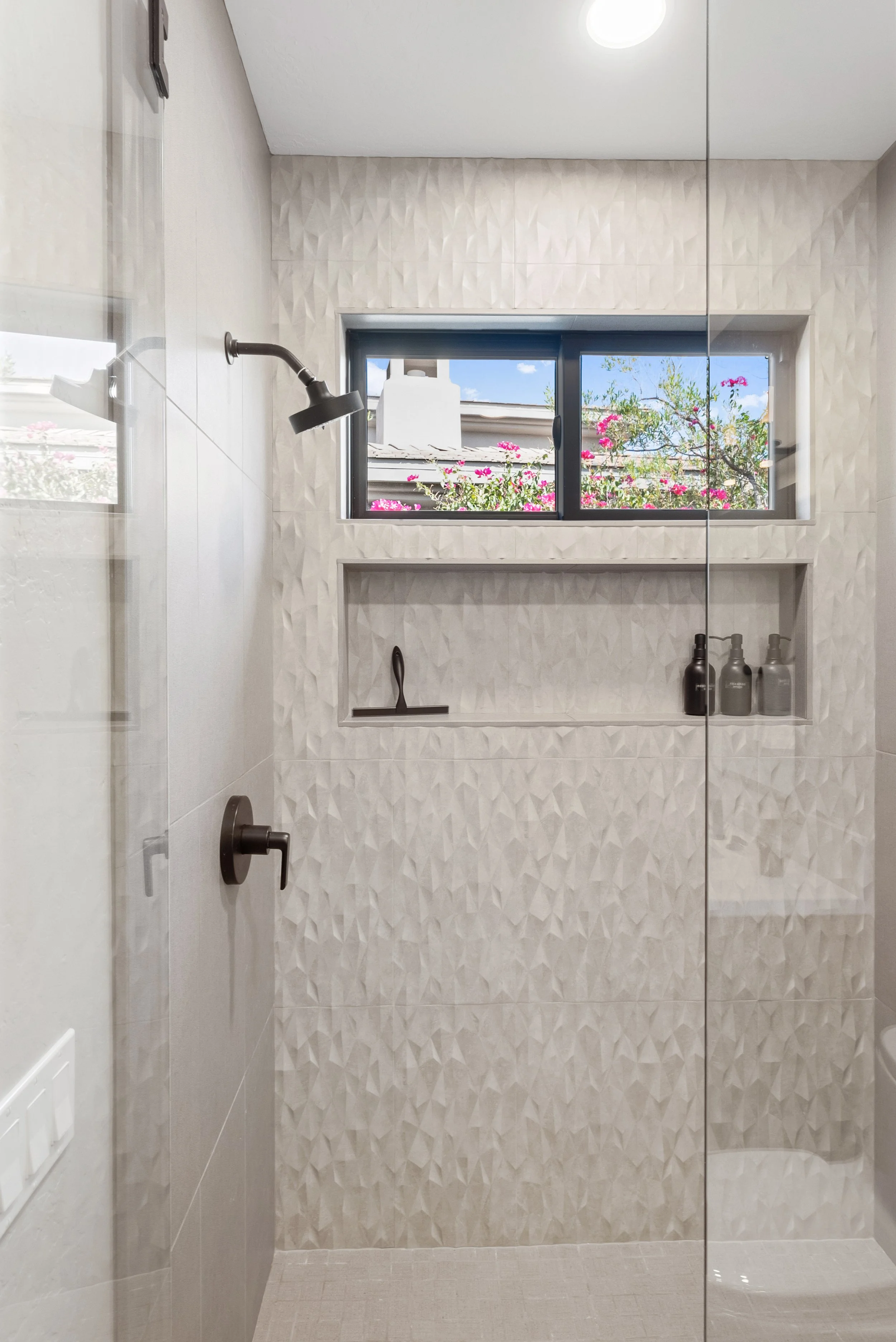 A modern shower with beige textured wall tiles, a black shower head, and a rectangular niche holding toiletries. There is a window outside with a view of pink flowers and blue sky.